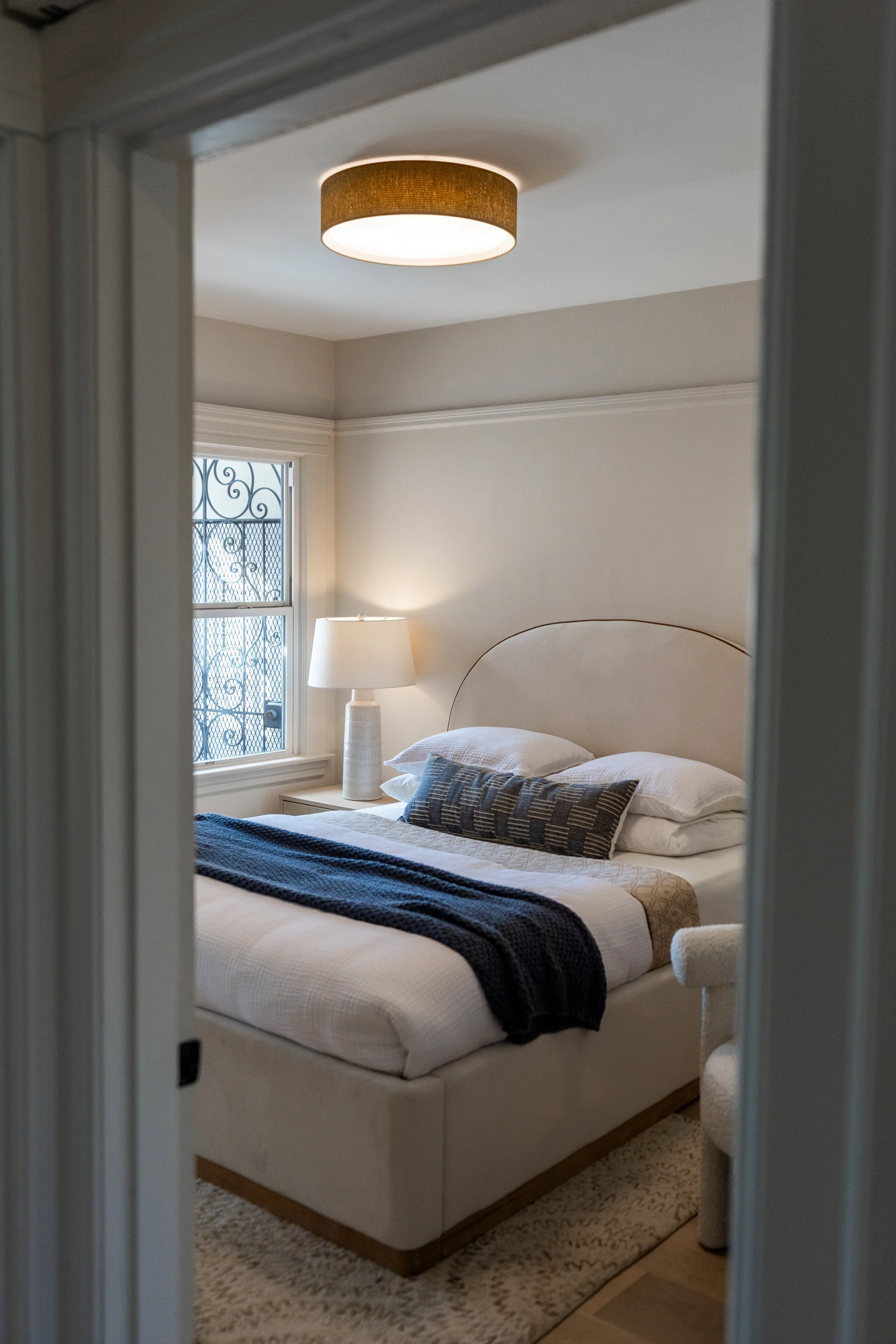 A bedroom with a bed that has white linens, black and white pillows, and a black throw blanket. There is a nightstand with a tall lamp next to a window with decorative ironwork. A ceiling light fixture is turned on, illuminating the room.