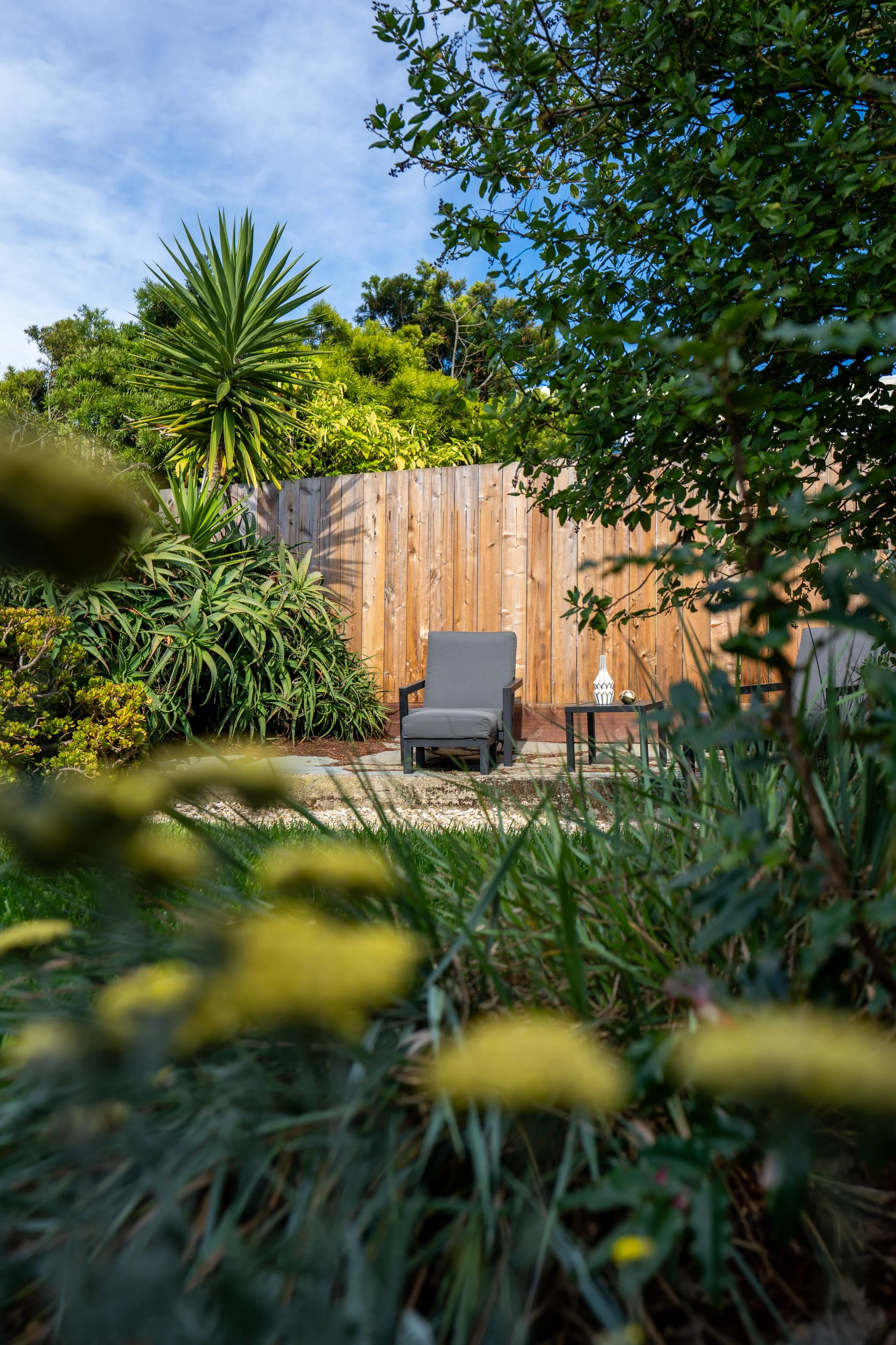 View of a backyard garden with a wooden fence, surrounded by lush green plants, including a large spiky plant, with a chair and side table with a decorative vase on top under a partly cloudy sky.