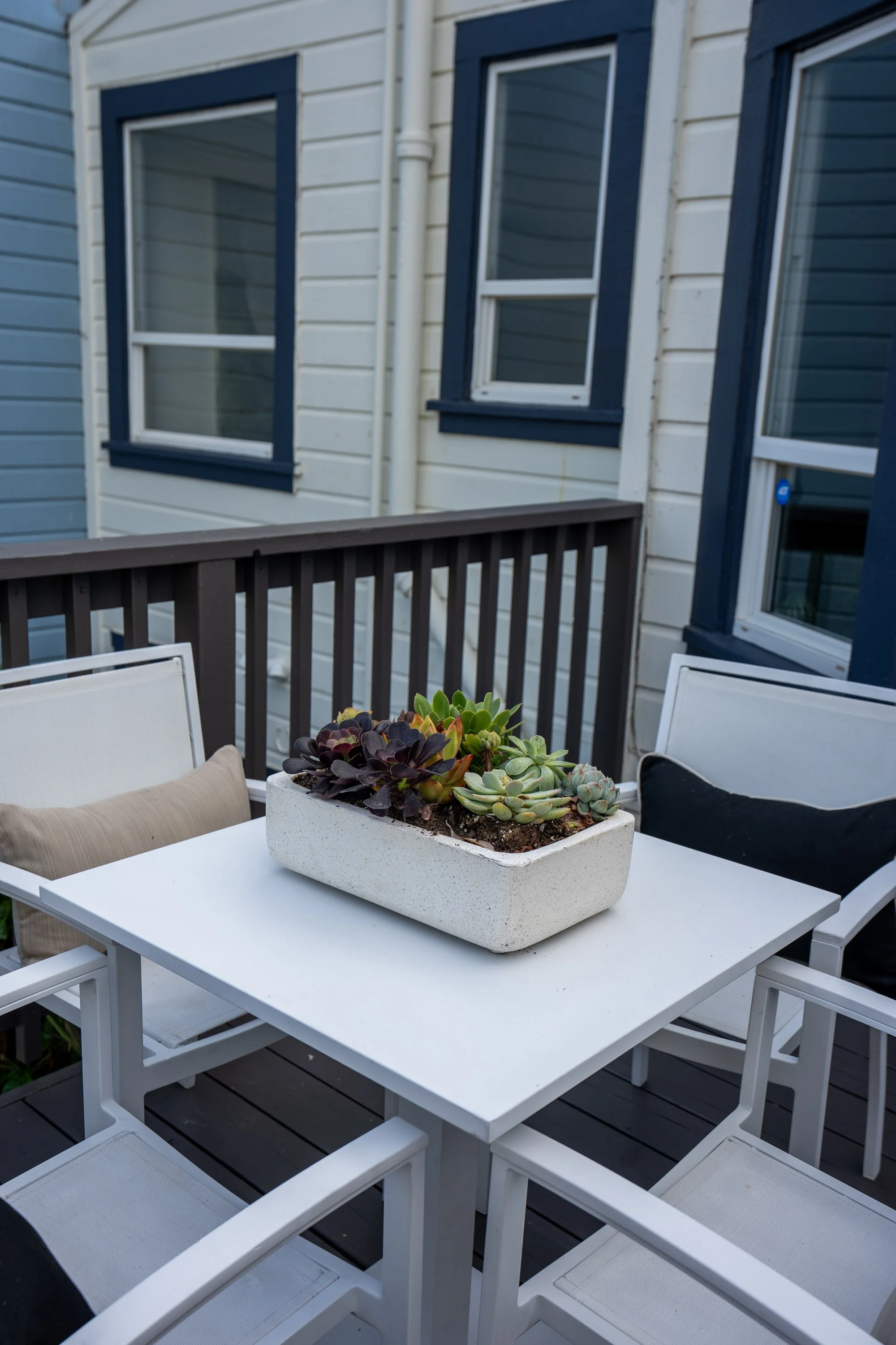 Outdoor patio with a white rectangular table, surrounded by white chairs, and a rectangular planter with succulents on the table. The patio has a wooden railing and a background of a house with white siding and blue window frames.