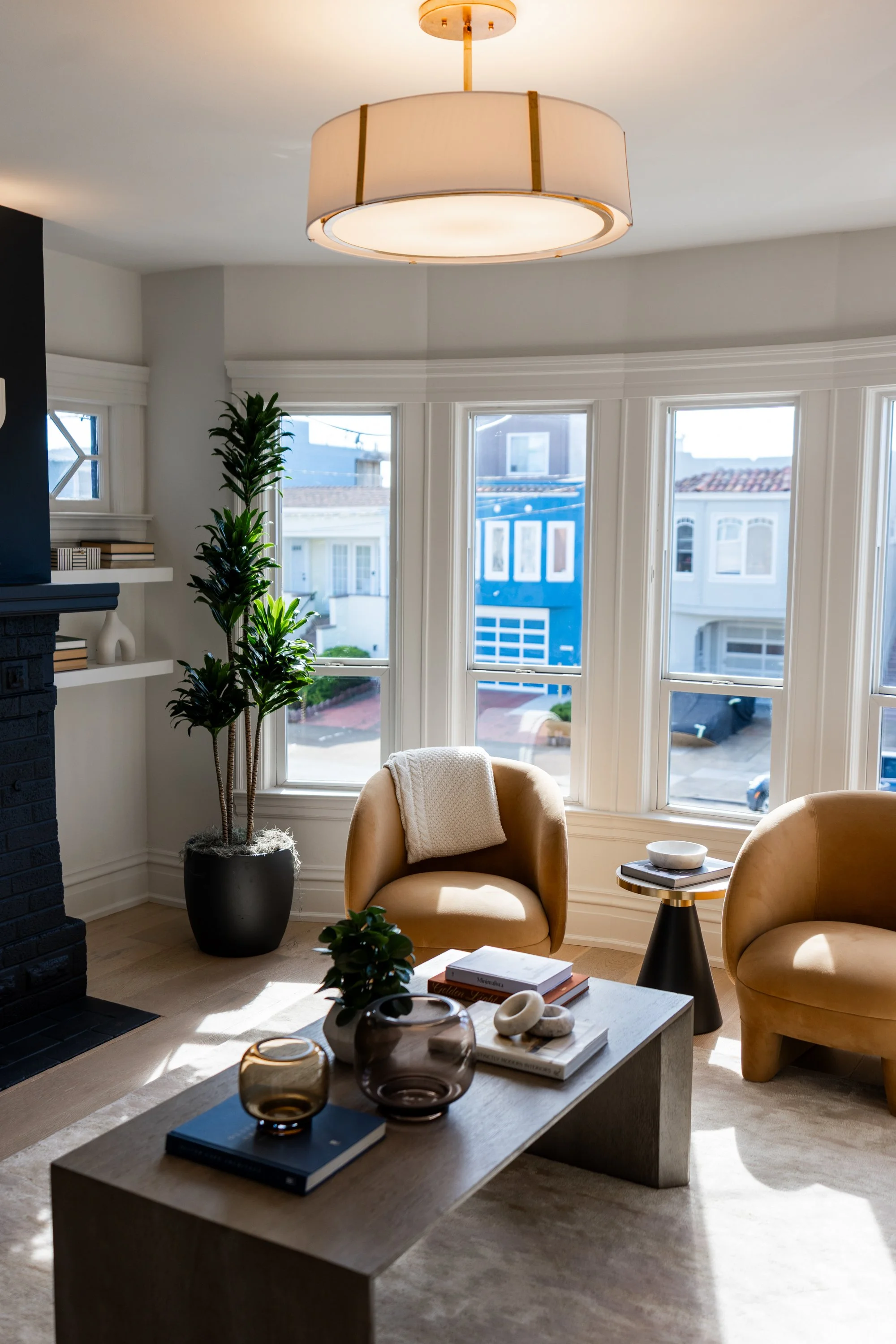 Living room with beige armchairs, a coffee table with books and decorative objects, a tall plant, large windows showing city houses, and a ceiling light fixture.