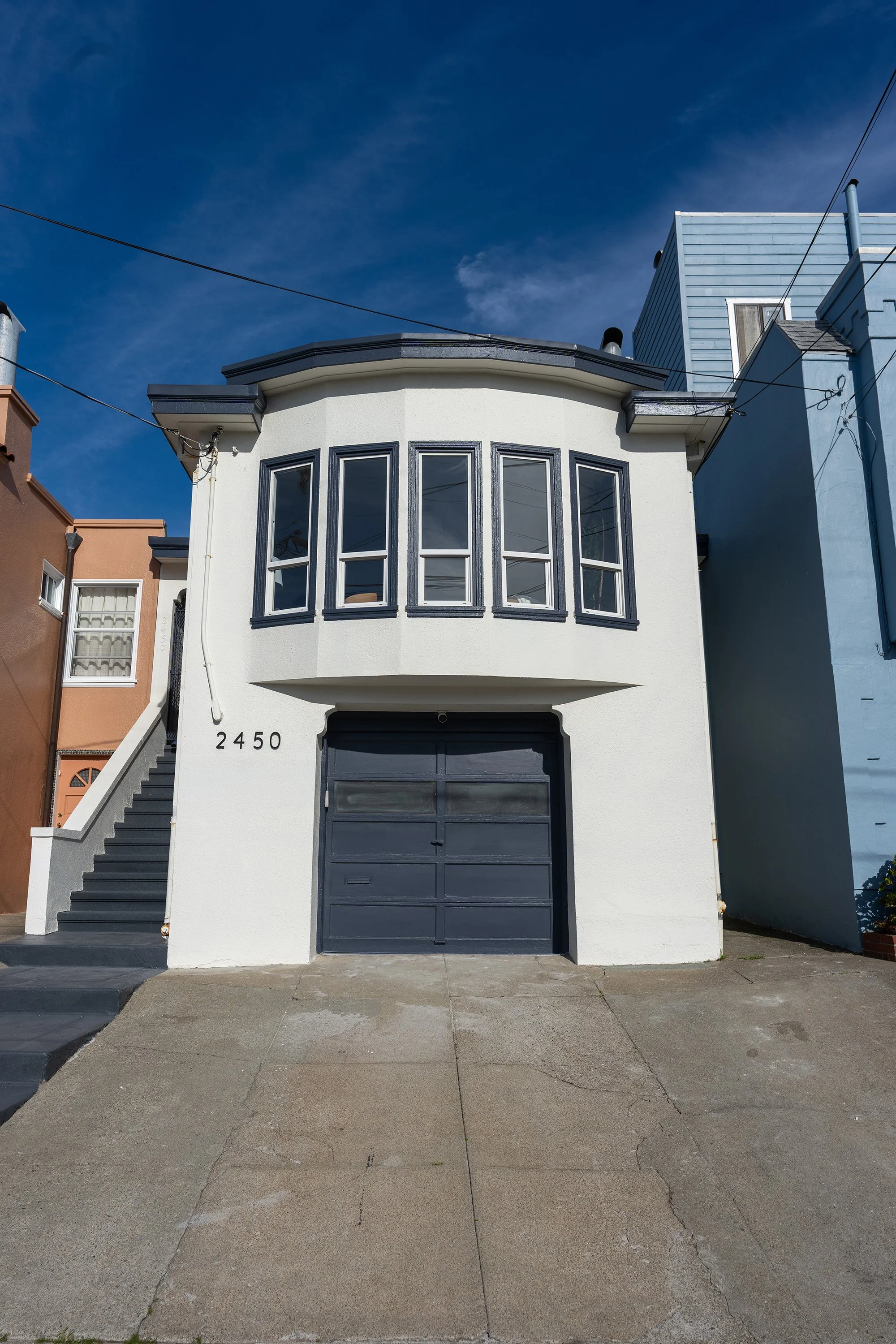Front view of a white two-story house with a black garage door and black trim around its windows. The house has stairs on the left side leading to an entrance, and the street in front is paved with concrete, showing some cracks. The house number is 2