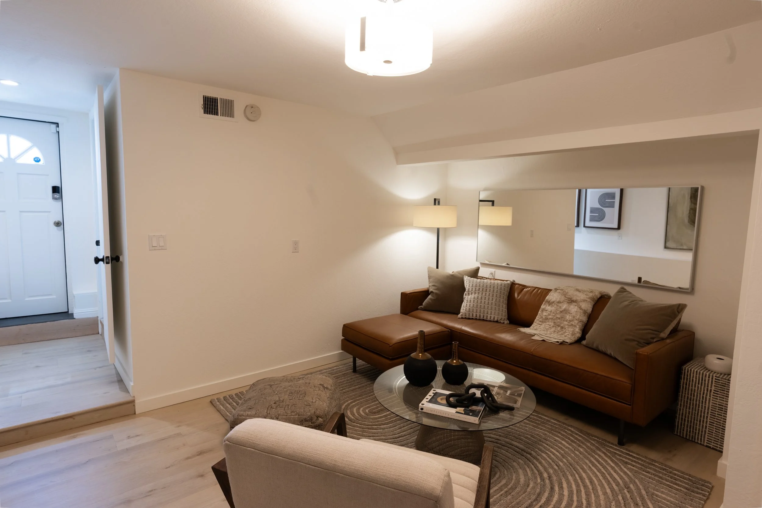 Living room with beige walls, a brown leather sofa with pillows, a glass coffee table with black vases and books, a light-colored rug, a floor lamp, a framed abstract artwork, and a large mirror on the wall.