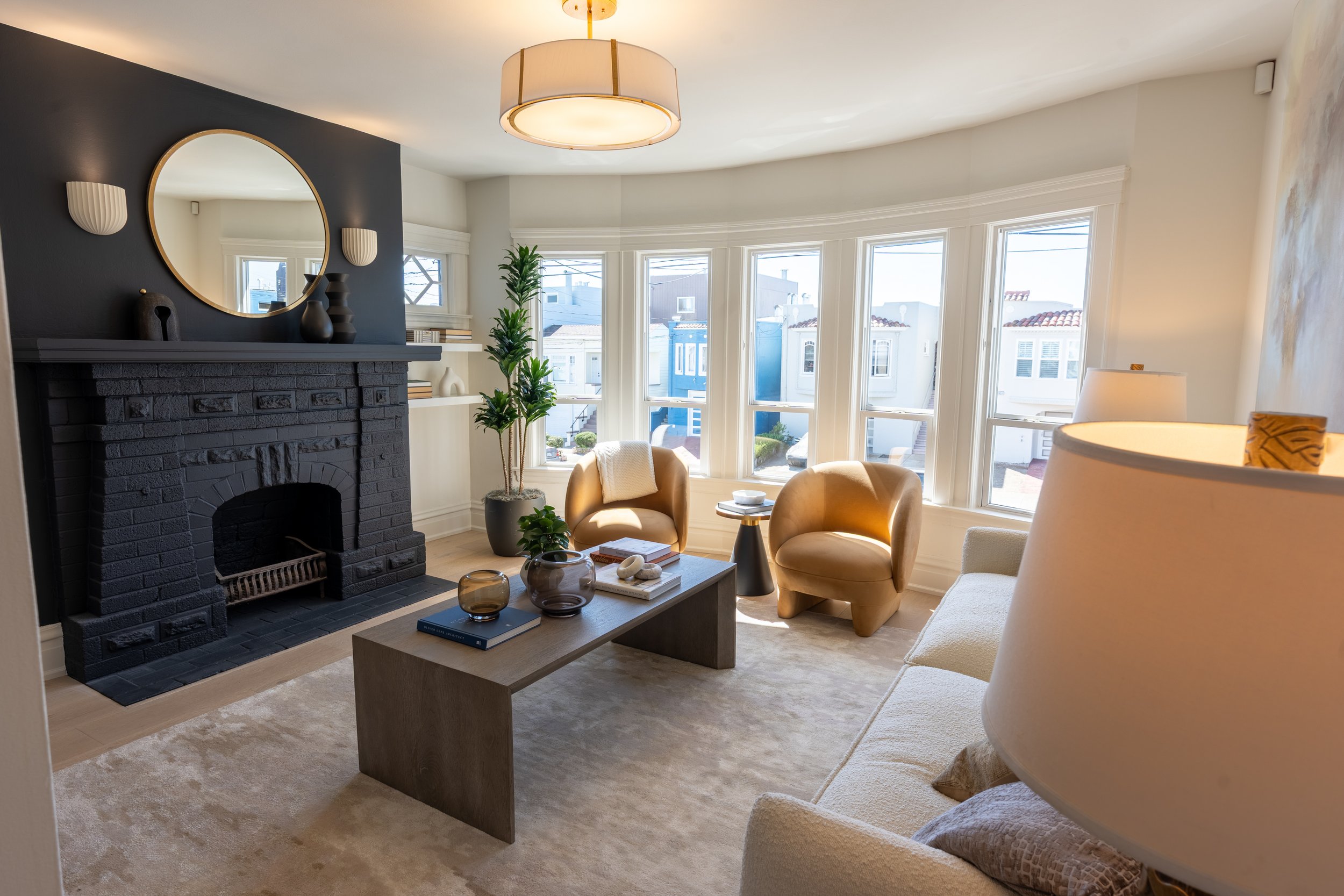 Living room with a fireplace, mirror, and large bay windows showing neighboring houses. There are two tan chairs, a coffee table with books and decorative items, and a tall potted plant.