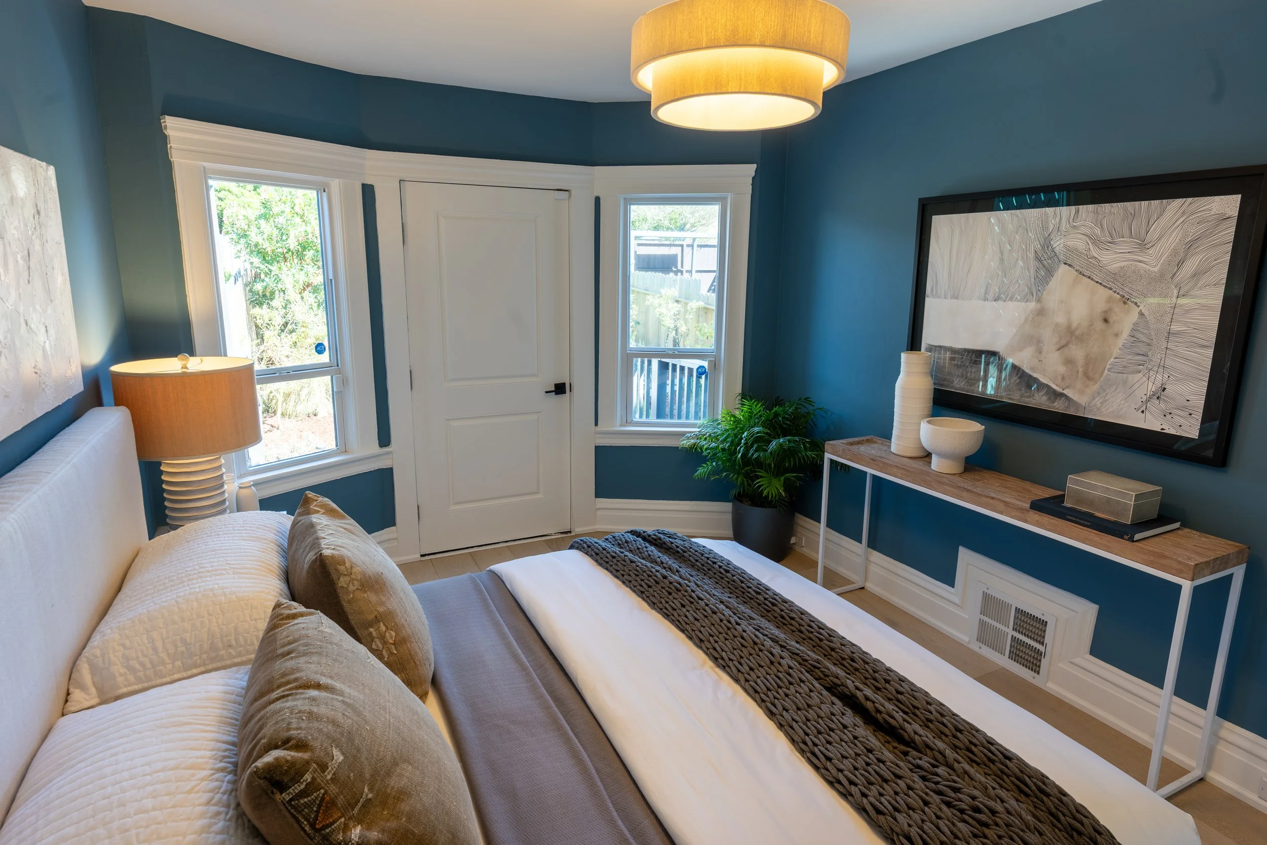 A cozy bedroom with blue walls, two windows, a white door, and a beige bed with pillows. The room has a brown bedside lamp, a green potted plant, a white console table with decorative vases, a large framed black and white artwork, and a beige ceiling