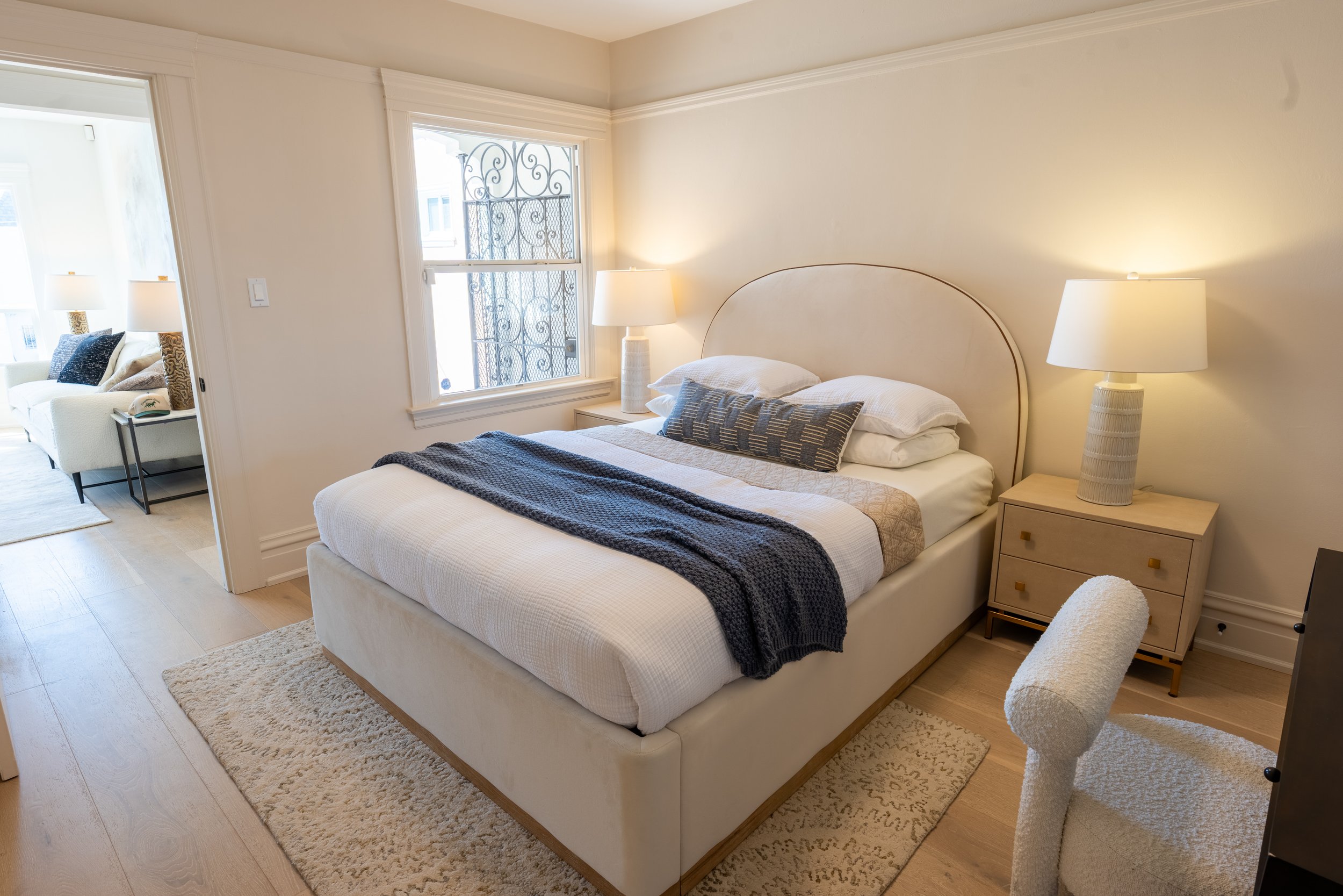 A bedroom with a bed, nightstand, and lamps, adjacent to a sitting area with a sofa and side table.