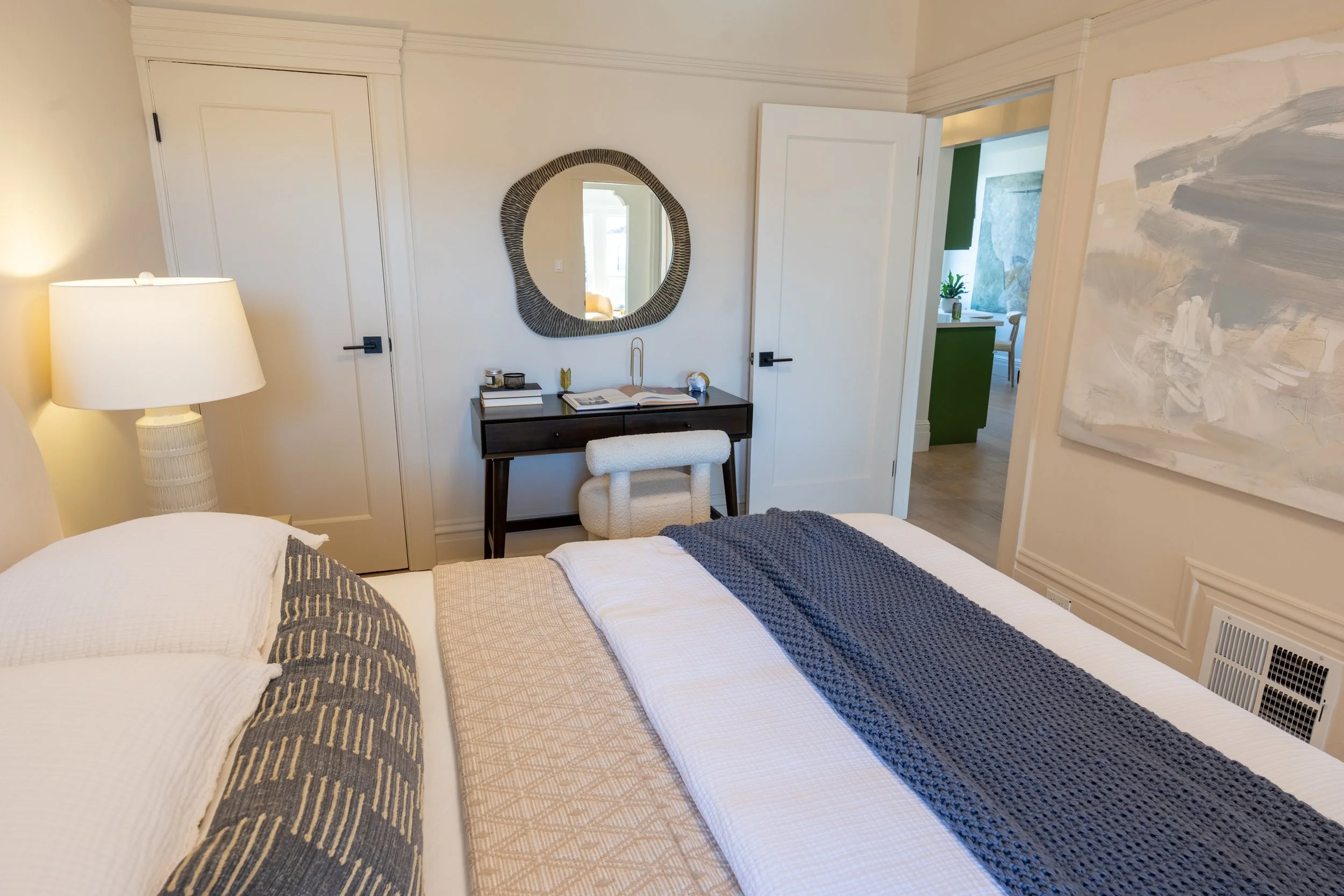 A cozy bedroom with a bed covered in a beige and navy blue quilt, a white bedside lamp, a small desk with books and decorative items, a round mirror, and an open door leading to another room with green accents and a window.