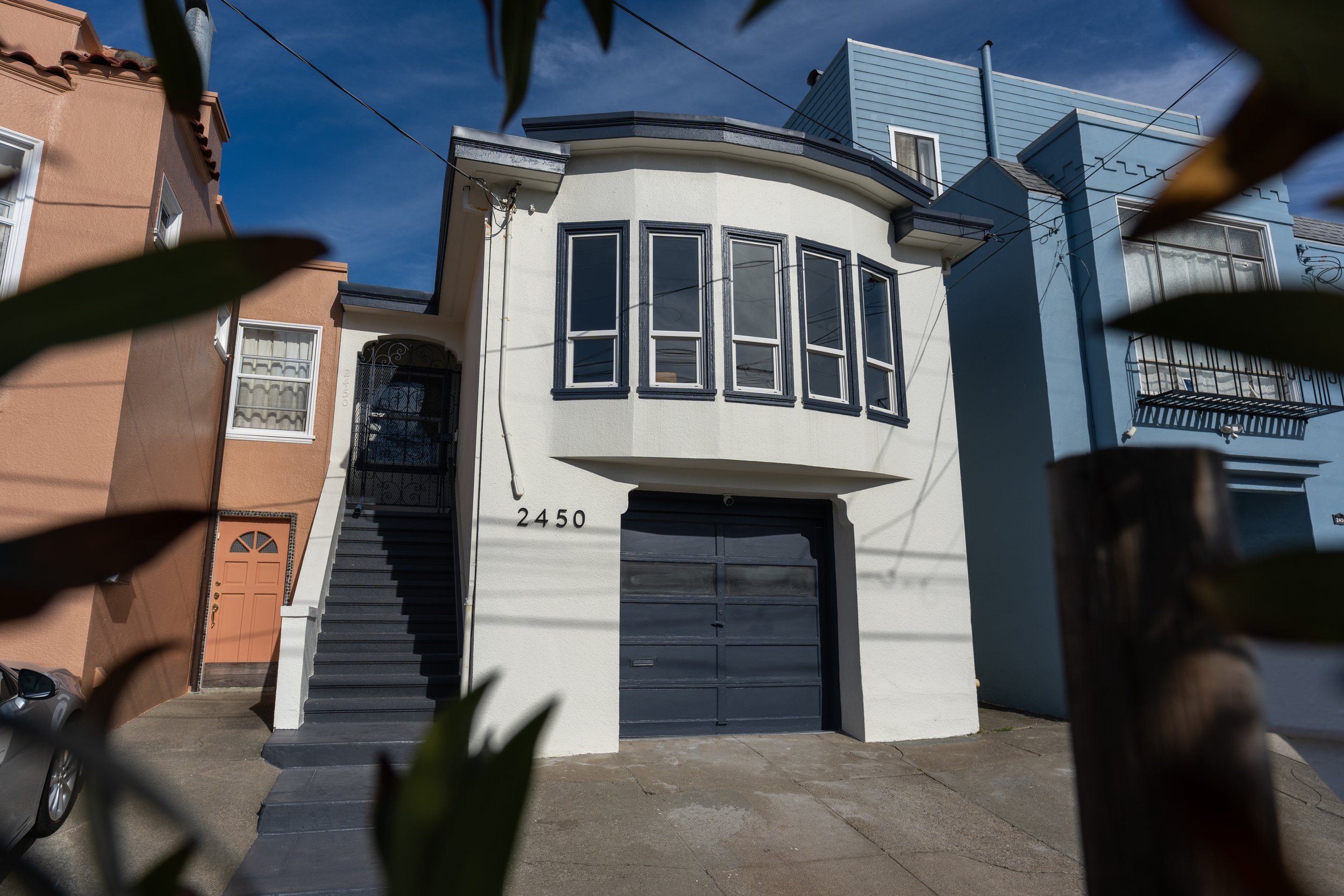 Front view of a modern multi-story house with white and blue exterior, bay windows, black garage door, concrete stairs, and house number 2450, framed by blurred leaves in the foreground.