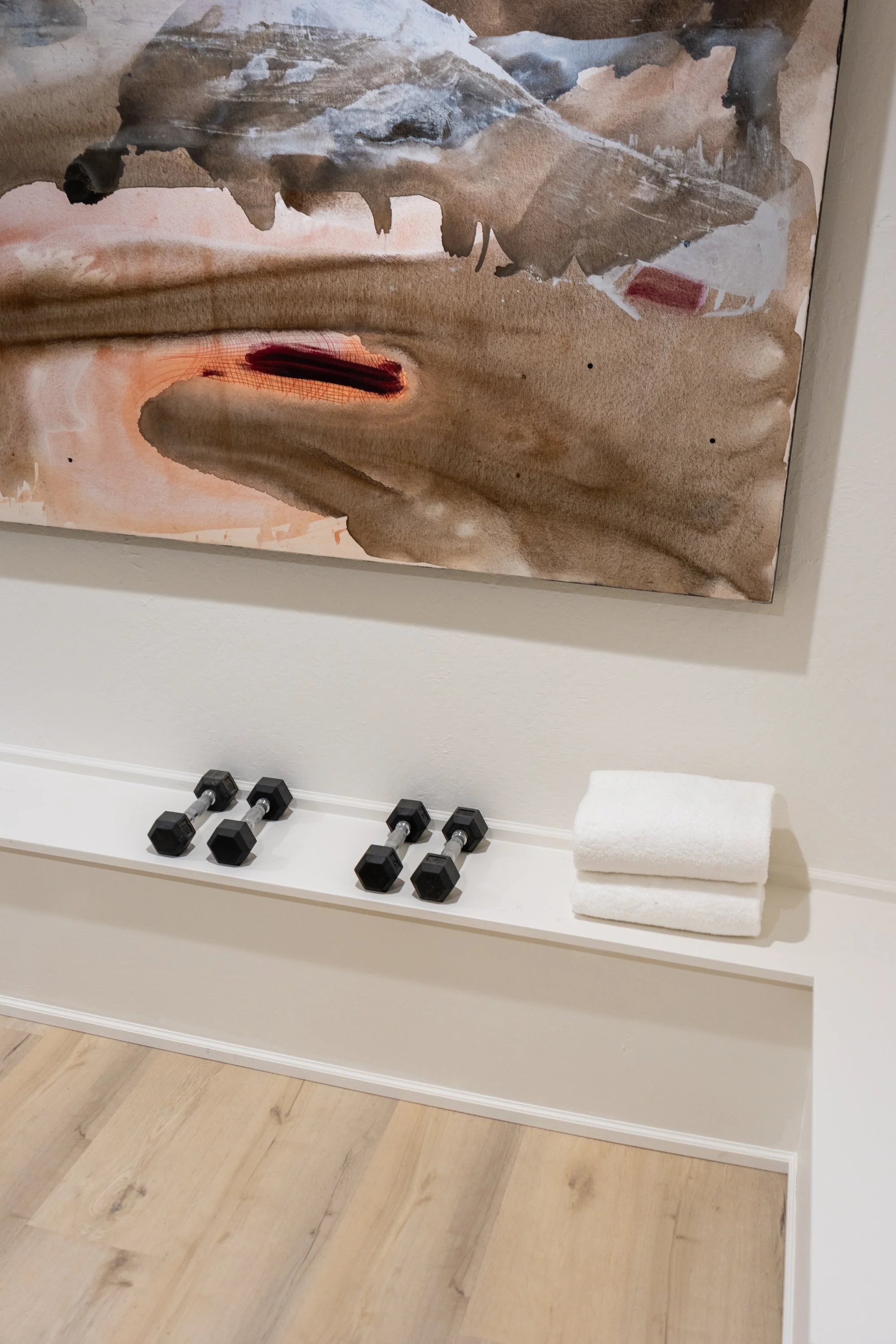 A white bench holds four black dumbbells, a stack of two white towels, and a rolled white towel. A large abstract watercolor painting hangs above the bench.
