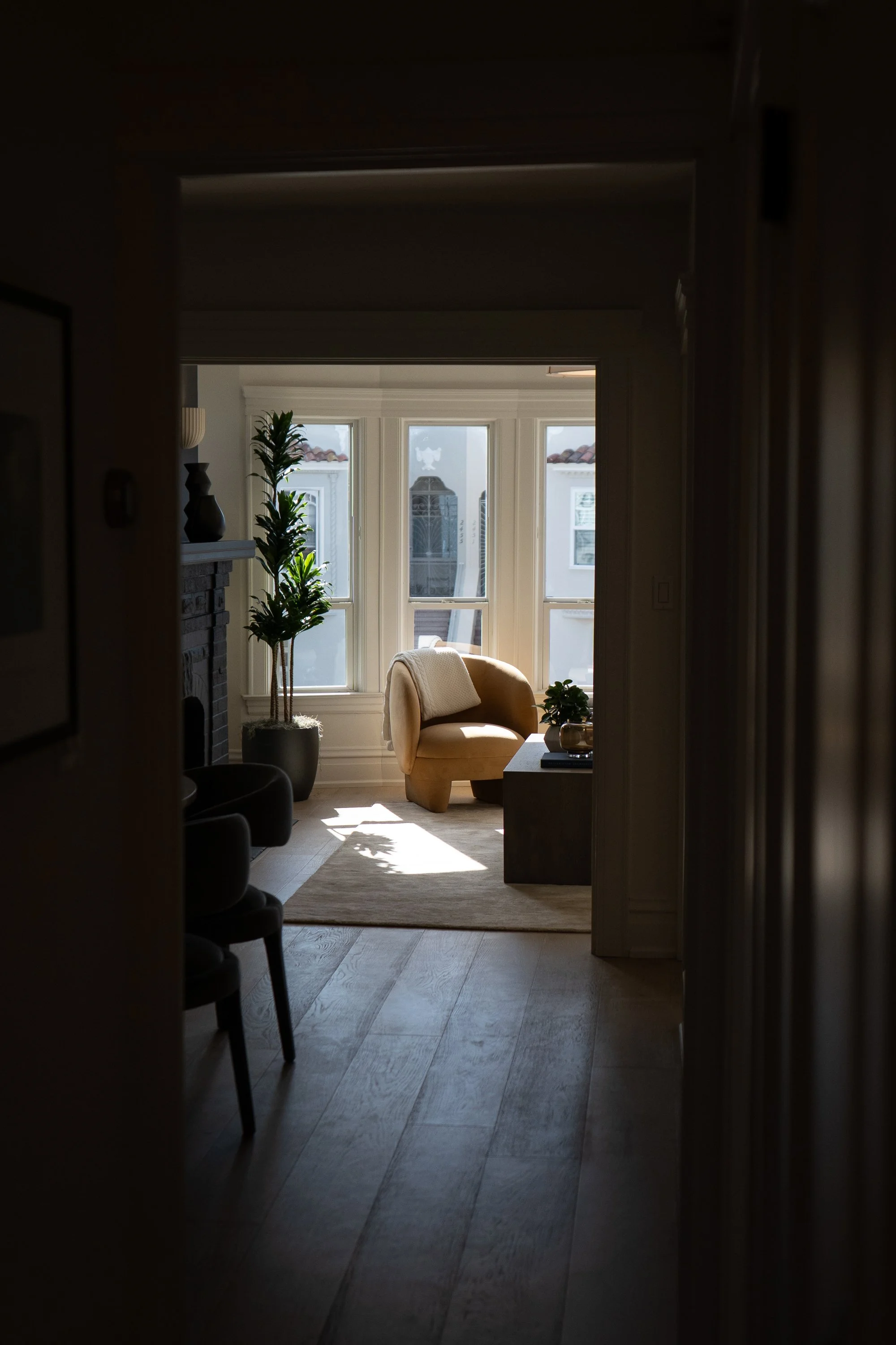Bright living room with large windows, beige armchair, potted plant, and decorative objects, viewed from a dim hallway.