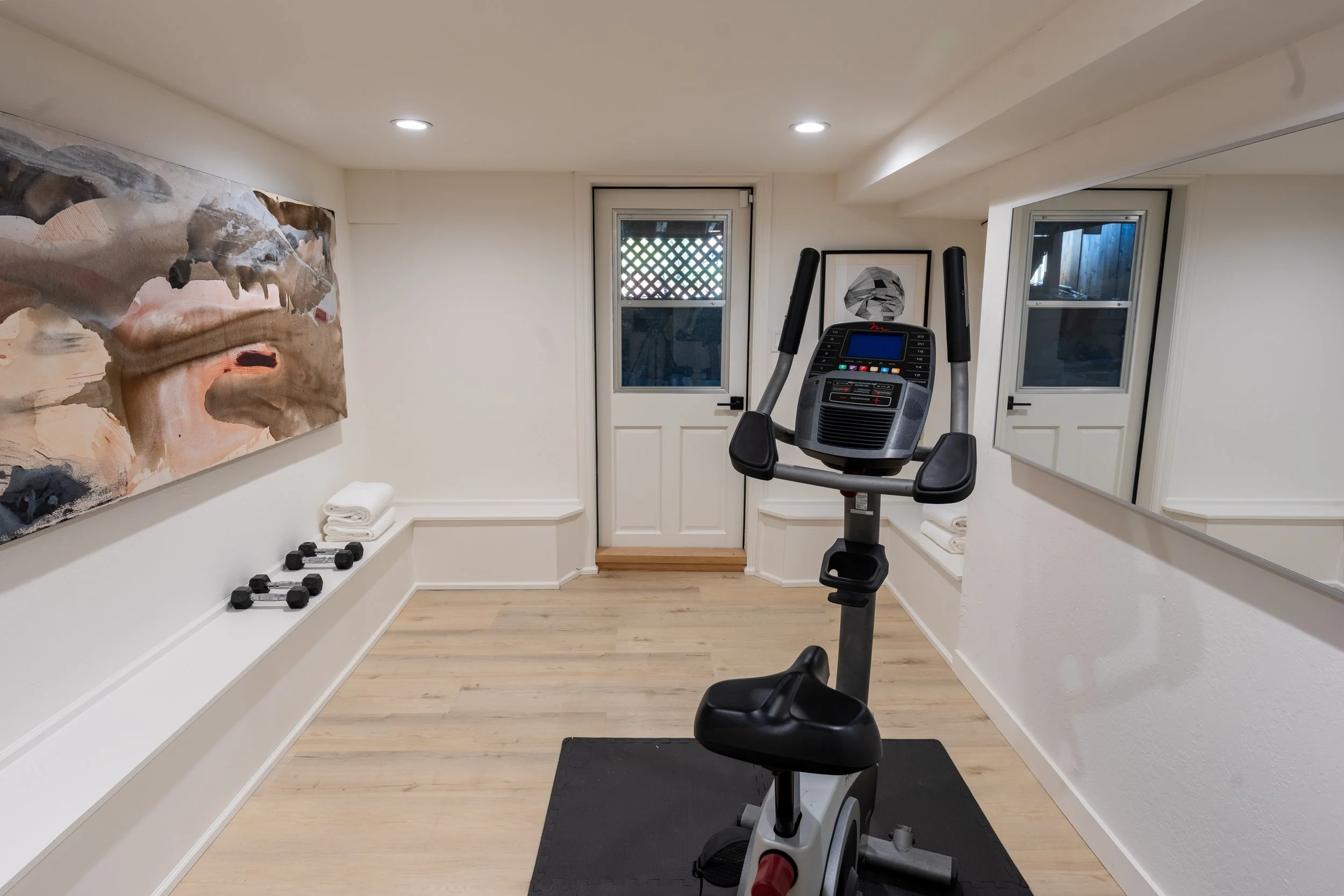 A basement workout room with an exercise bike, black mirror, abstract art, set of dumbbells, and a door leading outside.