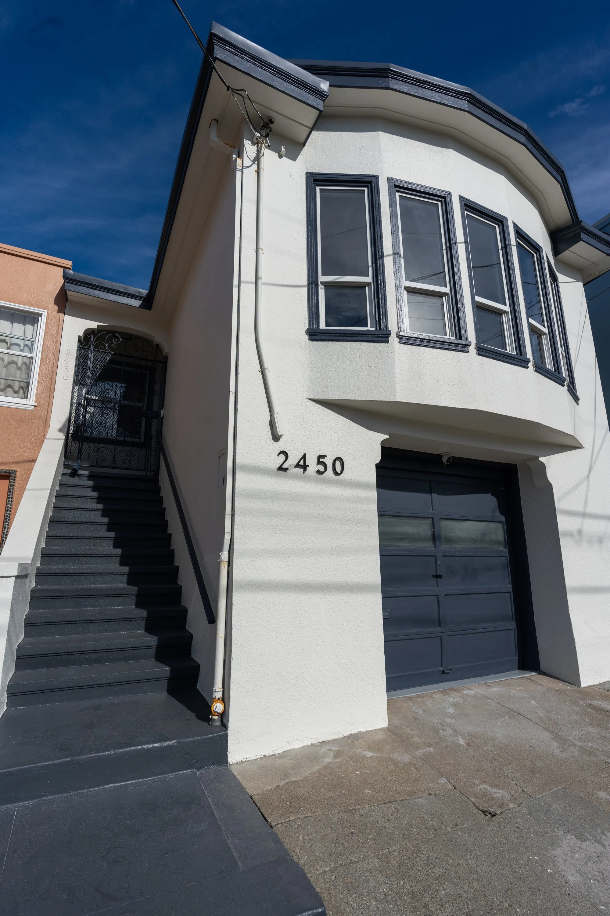 A modern white two-story building with black trim, large front windows, a garage door, and a black staircase with a wrought iron gate leading to the front door. House number 2450 is visible.