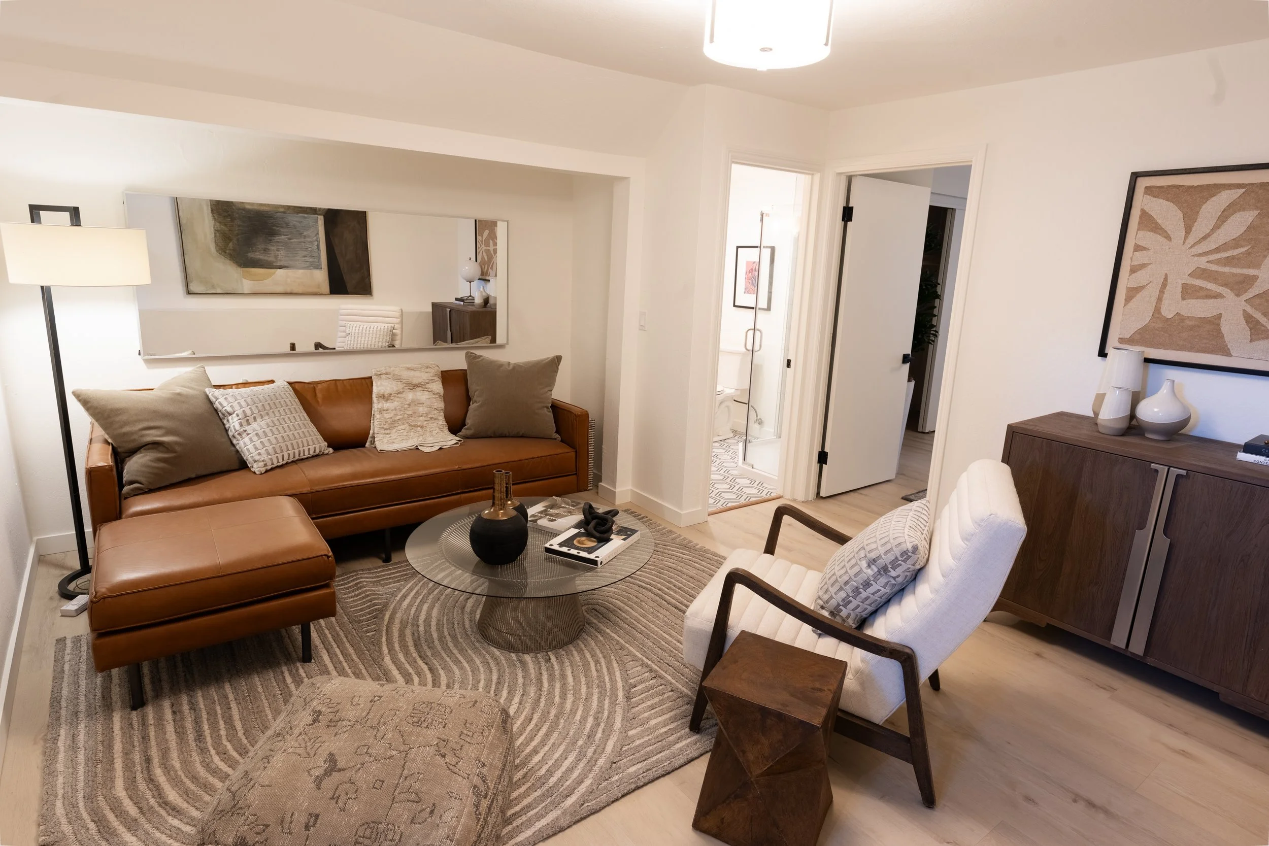 Living room with tan leather sofa, white armchair, wooden furniture, artwork on walls, mirror, lamps, decorative objects, and a view into a bathroom with black and white patterned floor.