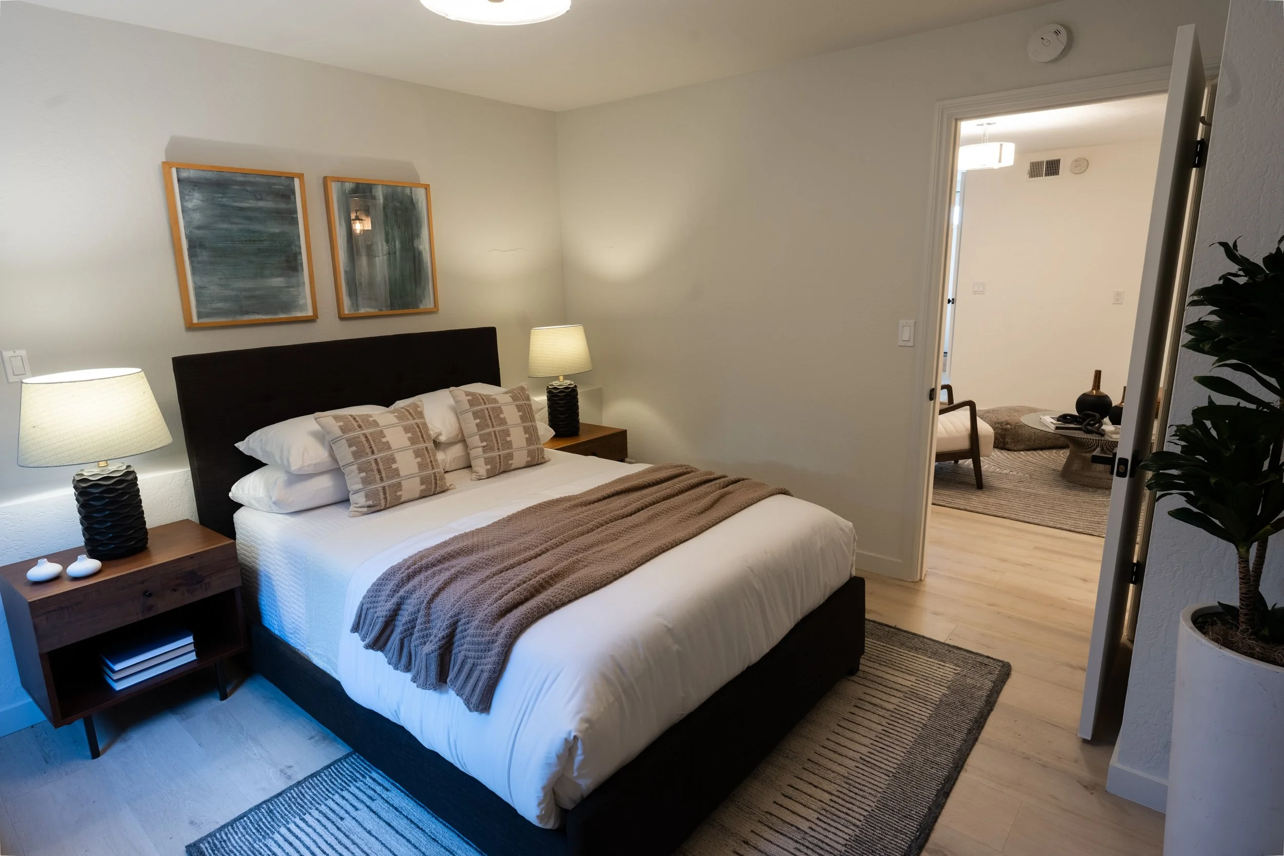 A bedroom with a bed, two lamps, abstract artwork, and a nearby sitting area visible through an open door.