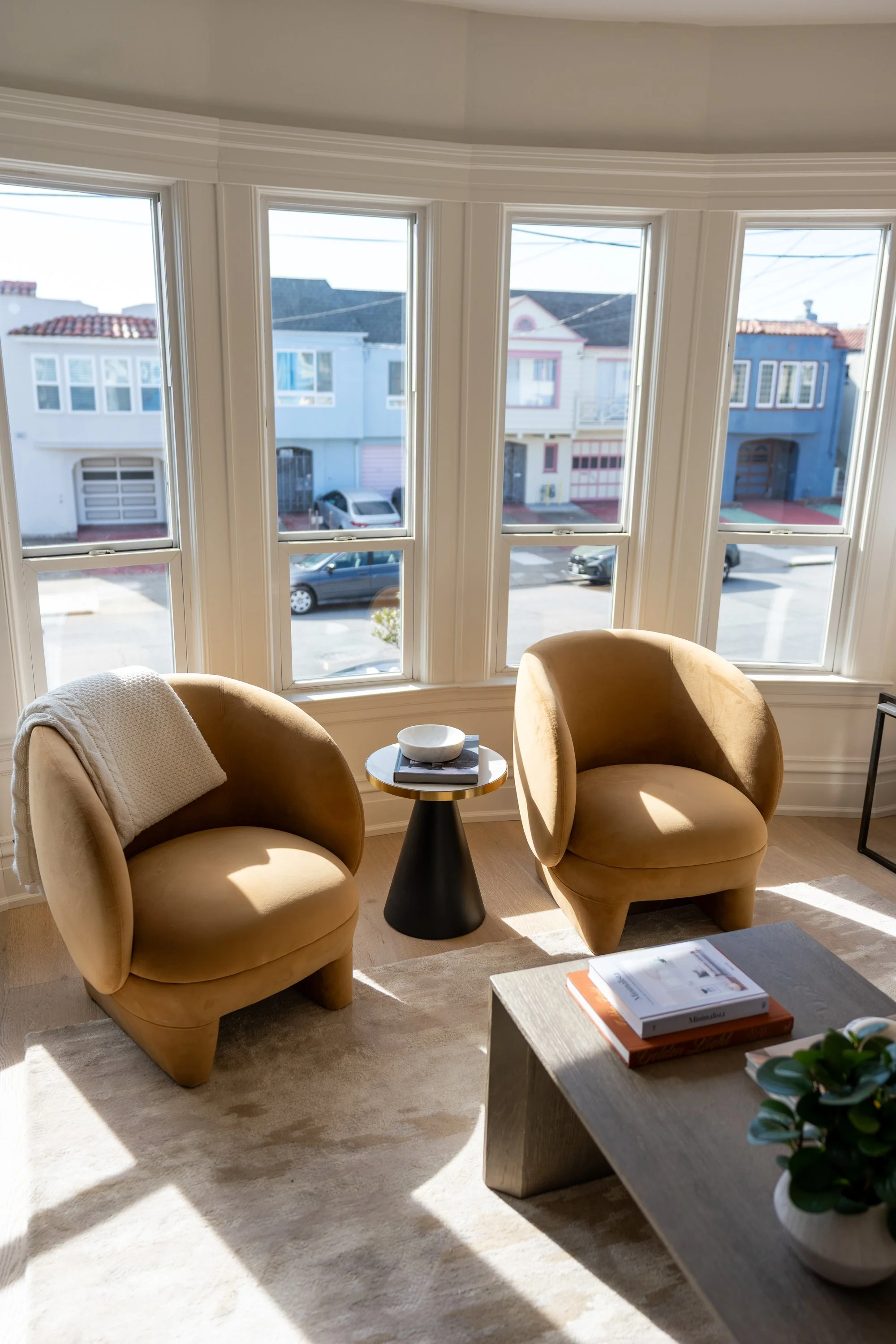 A cozy living room corner with two beige armchairs, a small round side table with a bowl and books, a potted plant on a nearby table, large bay windows showing a street with parked cars and colorful houses, and sunlight illuminating the space.