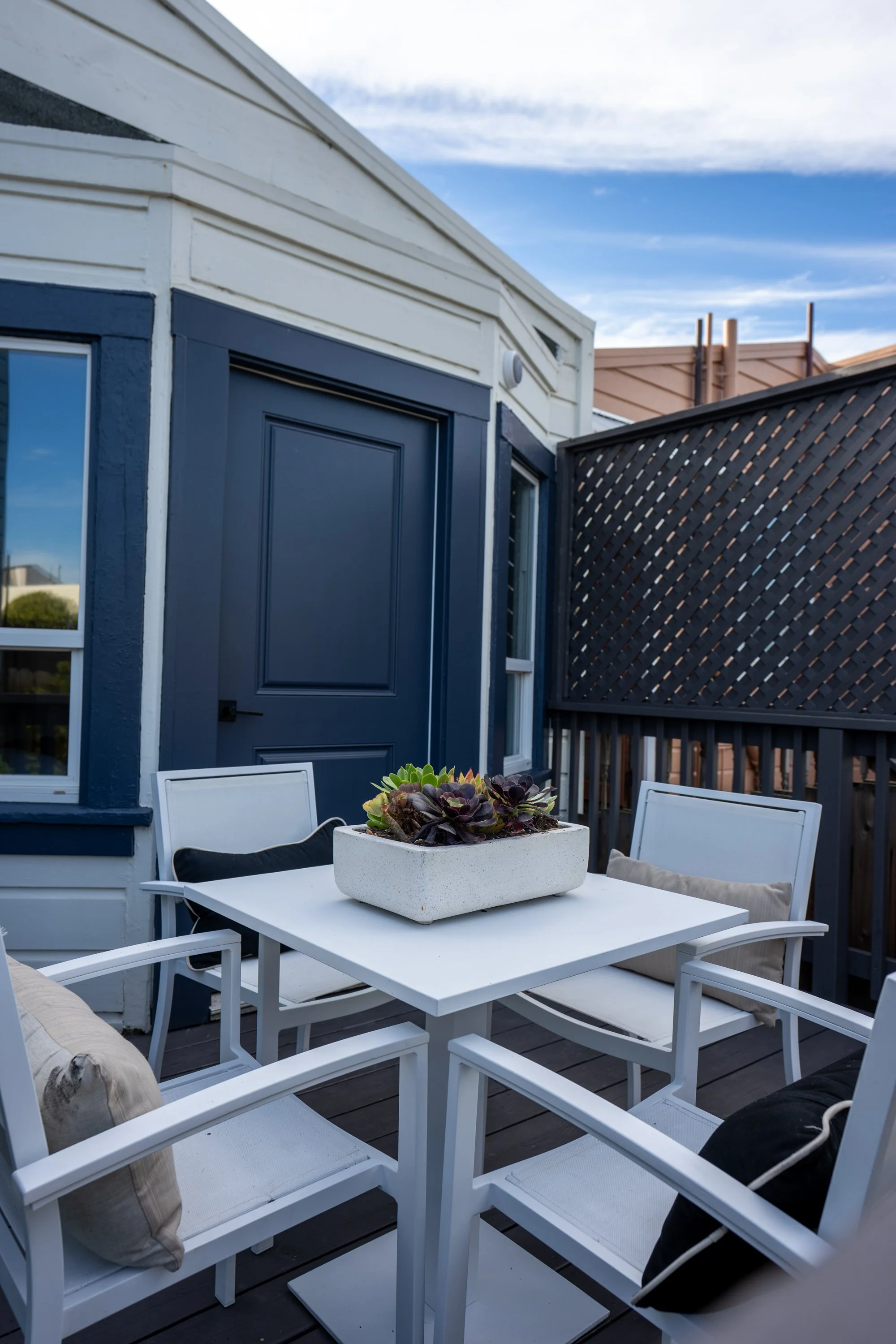 View of a rooftop patio with a white dining table and chairs, decorative pillows, a planter with succulents, dark blue painted building wall, window, and a black lattice privacy screen against a partly cloudy sky.