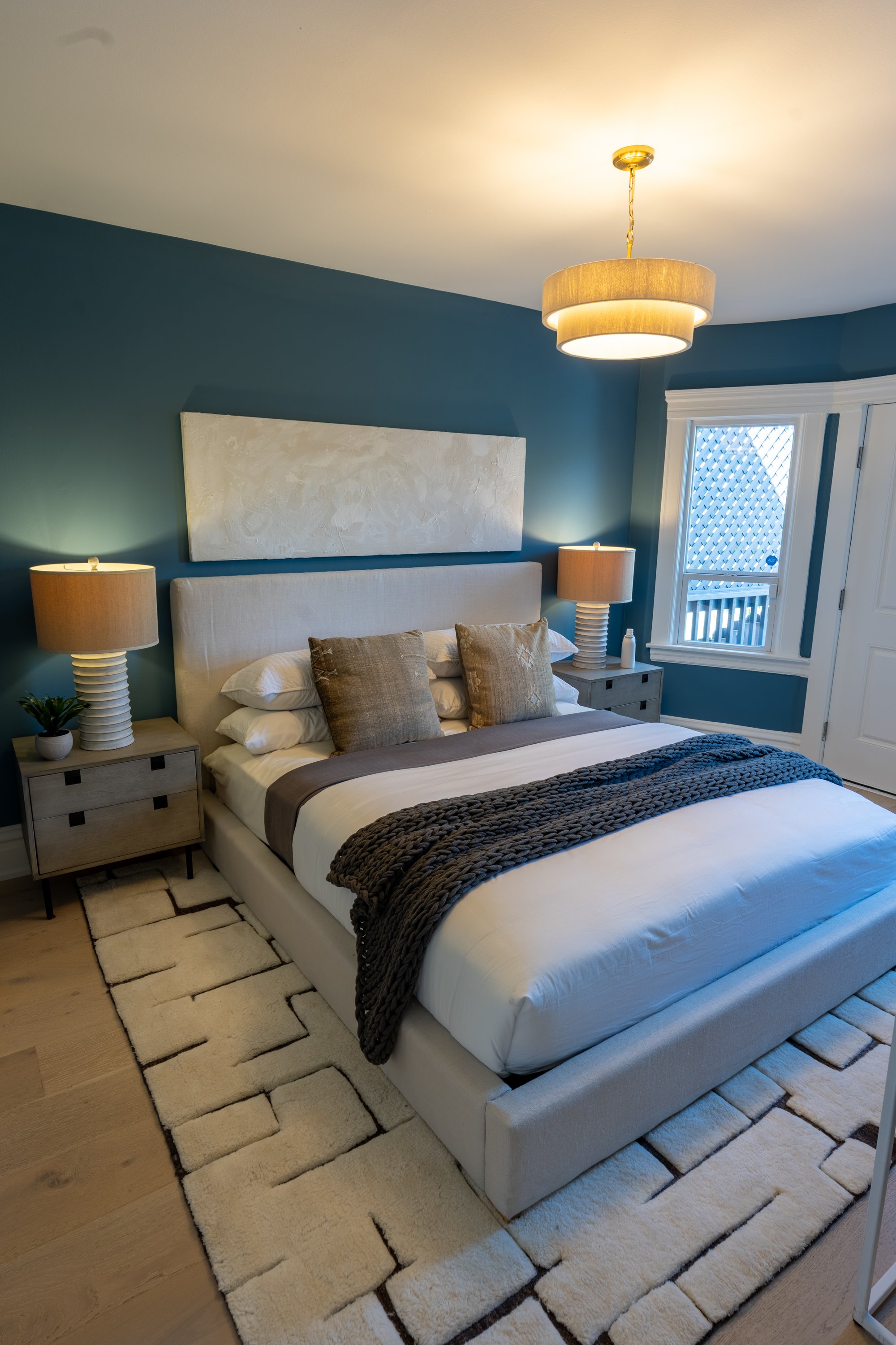 Bedroom with blue walls, beige upholstered bed with white bedding, gray and brown pillows, dark chunky knit blanket, two matching bedside tables with beige lamps, window with white frame and lattice, contemporary ceiling light fixture, light-colored 