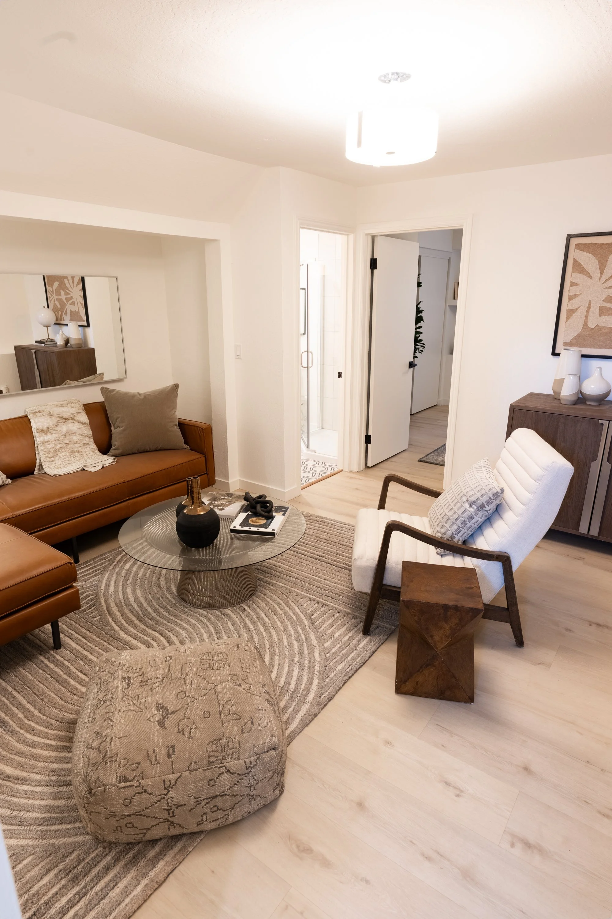 Living room with tan leather couch, white armchair with cushion, round glass coffee table with decorative items, area rug, sideboard with vases, and wall art. Open door leads to hallway or bathroom in a modern home.