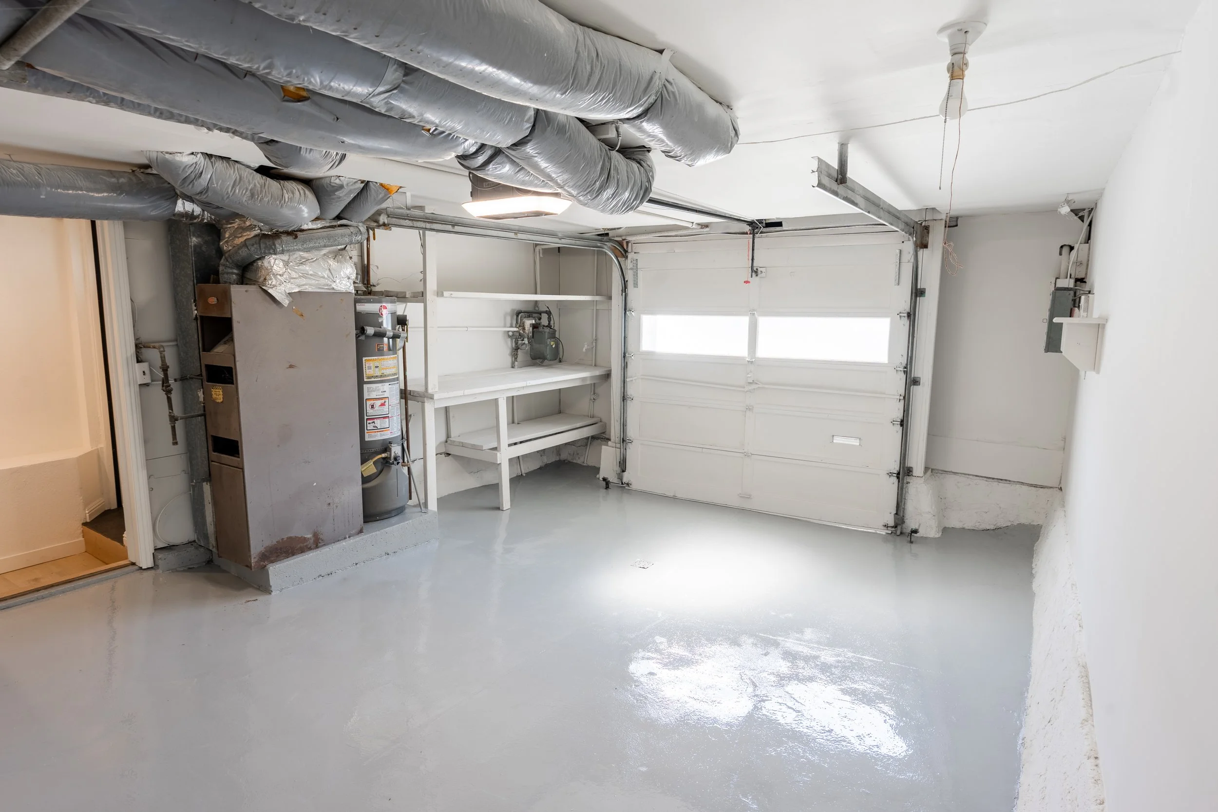 Empty garage with a clean, painted concrete floor, a closed white garage door, and exposed ductwork with insulation on the ceiling. There is a water heater and a small white shelf on the left side.