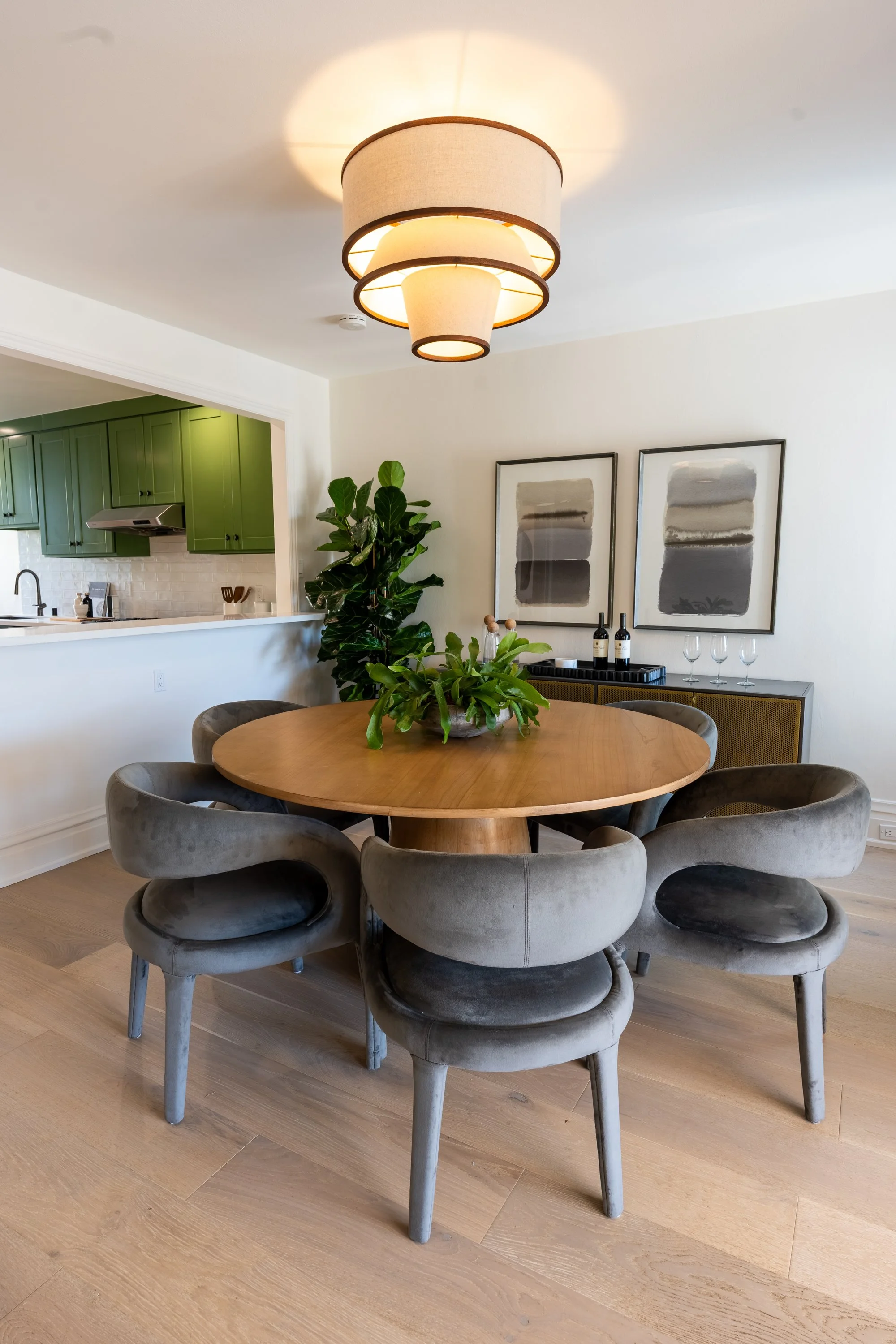 A round wooden dining table with six gray velvet upholstered chairs arranged around it, a large green leafy houseplant in the center, and a modern chandelier overhead. In the background, there is a kitchen with green cabinets and a white tiled backsp