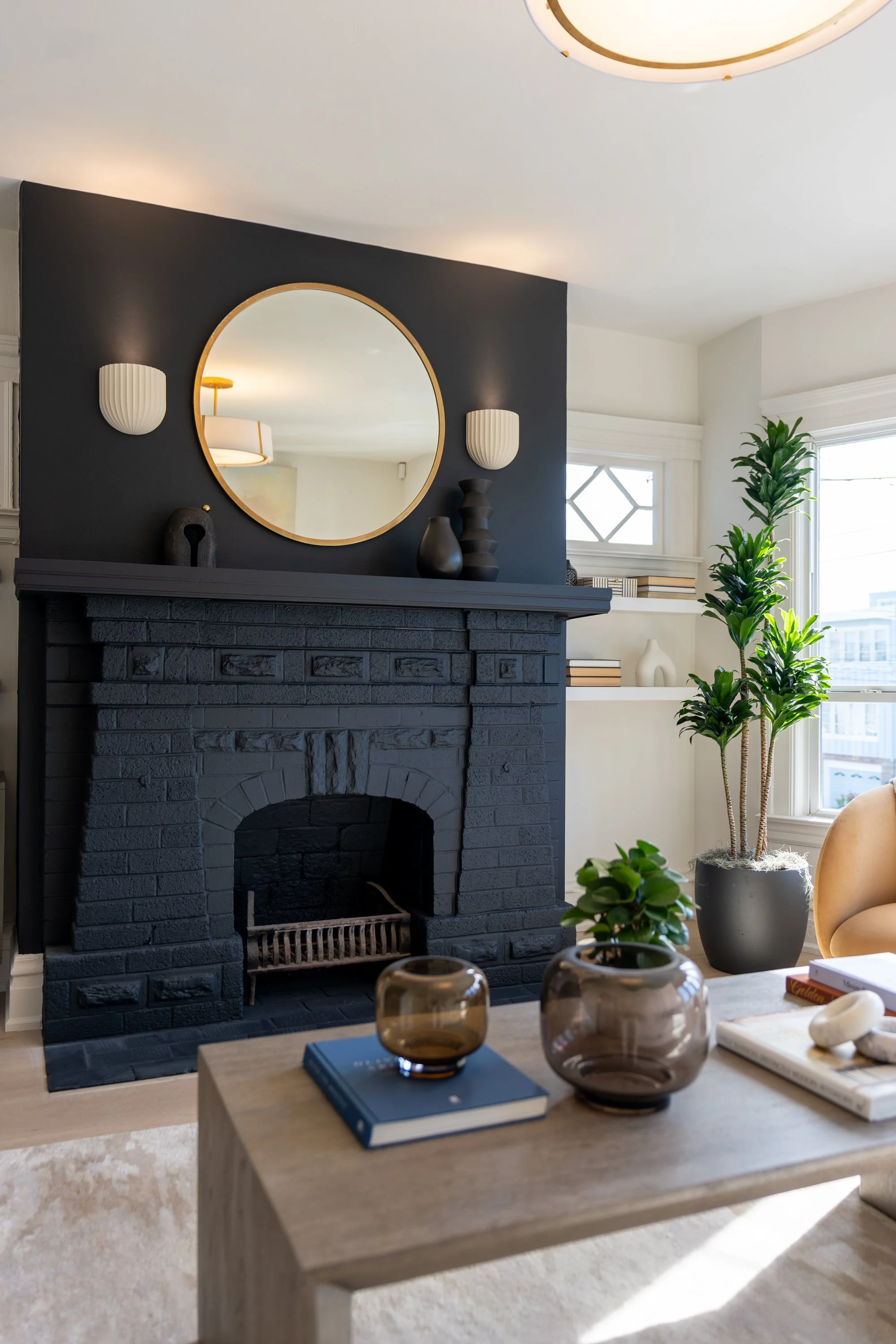 Living room with a black brick fireplace, circular mirror on dark accent wall, white wall sconces, and a wooden coffee table decorated with books and brown glass vases; presence of green plants near large windows with white trim, and a beige armchair