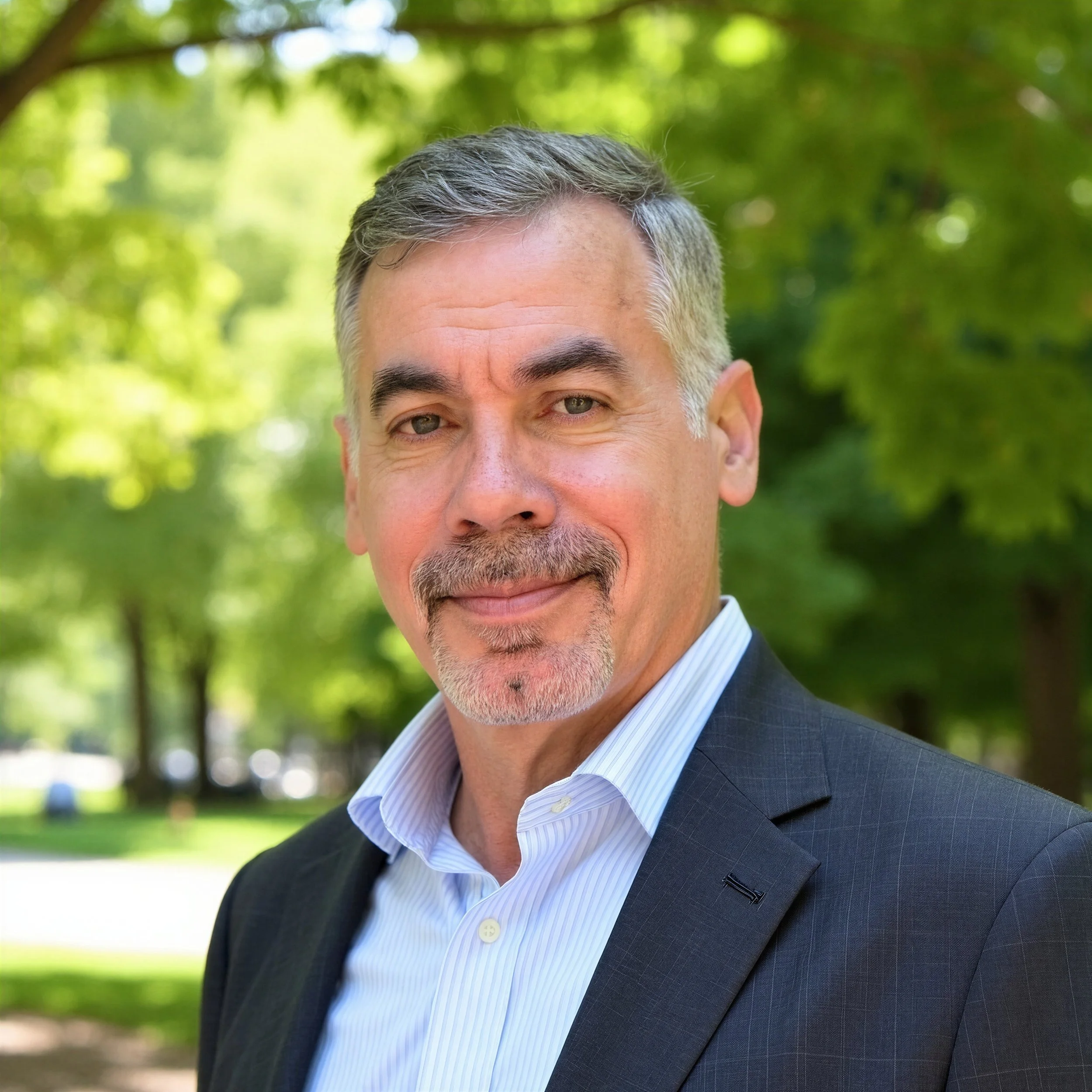 Close-up of a middle-aged man with gray hair and green eyes, wearing a dark suit and light blue striped dress shirt, smiling outdoors with a blurred green leafy background.