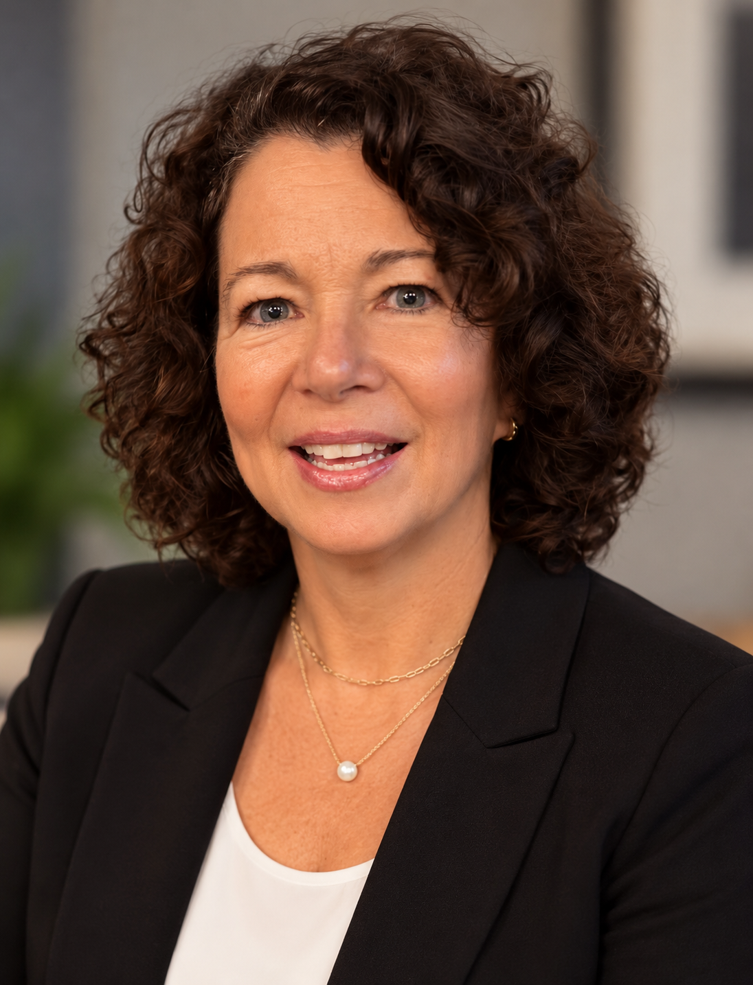 A woman with curly brunette hair smiling, wearing a black blazer and a pearl necklace, in an indoor office setting.