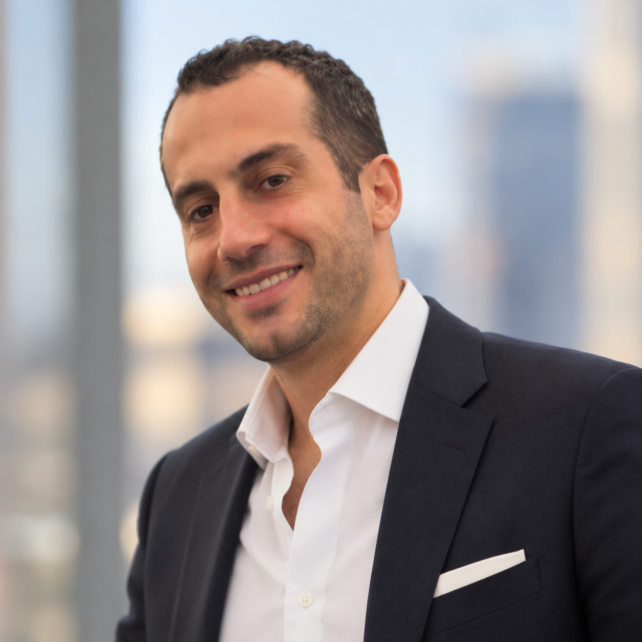 Headshot of a smiling man in a suit, standing in front of a blurred cityscape background.