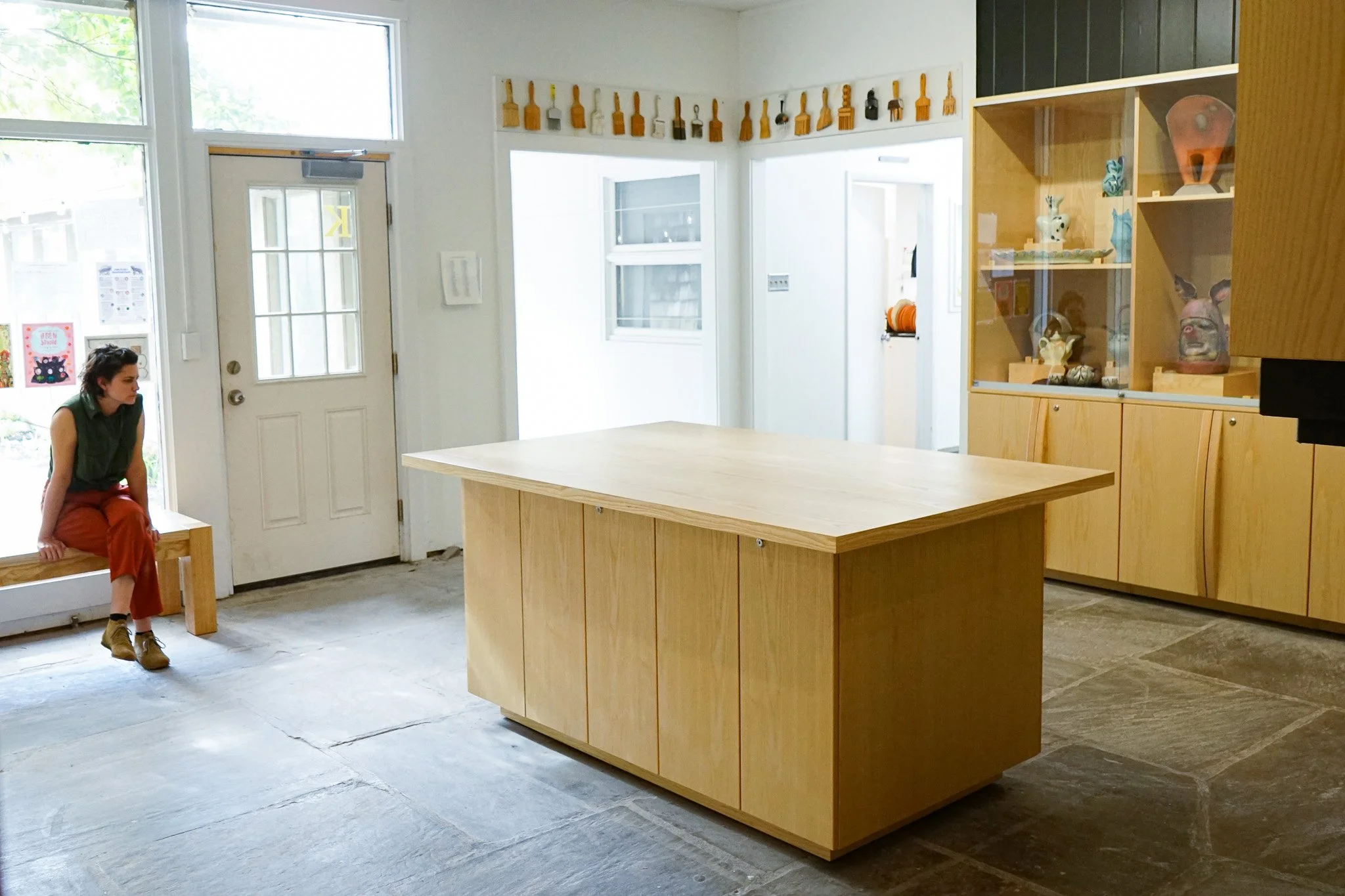 A woman sits on a wooden bench inside a room with white walls and large windows. There is a large, light wood kitchen island in the center and a wooden display case with decorative items on the right. The room has an industrial, modern aesthetic with