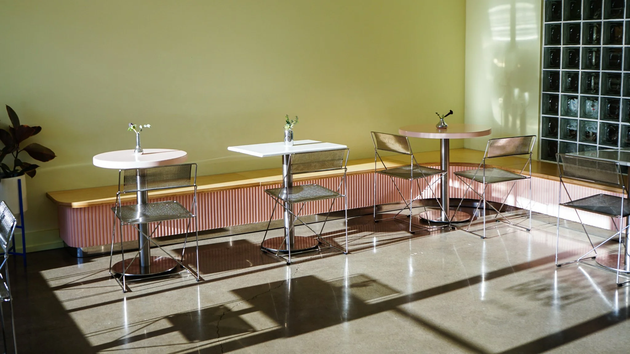 Empty cafe corner with three small tables, metallic chairs, a pink radiator cover, a yellow wall, and a glass block window with sunlight casting shadows.