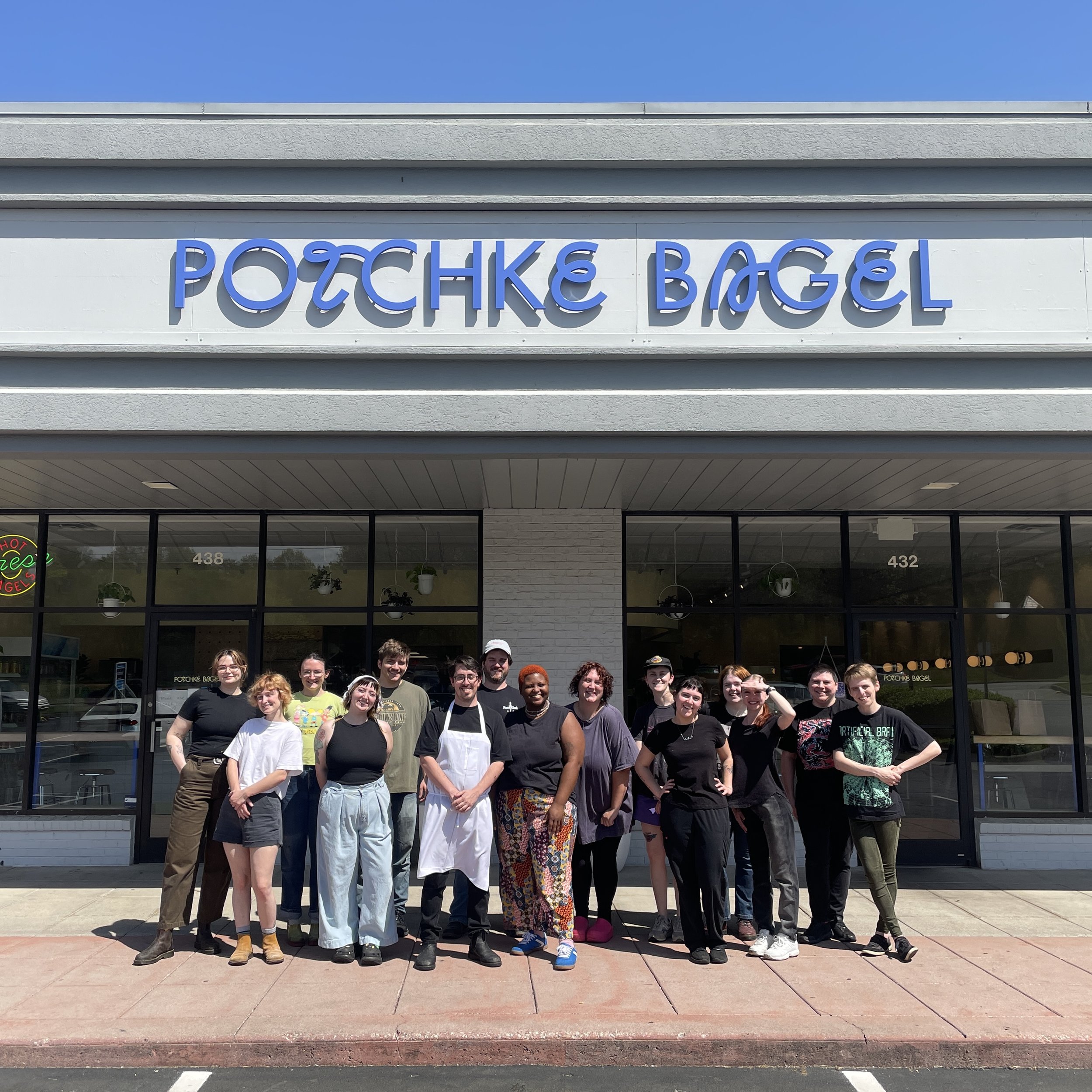 A group of diverse people standing in front of a bagel shop called 'Potiche Bagel'. The storefront has large glass windows, with some customers inside, and a neon sign that says 'Open' is visible. The group is smiling and appears to be happy, with so