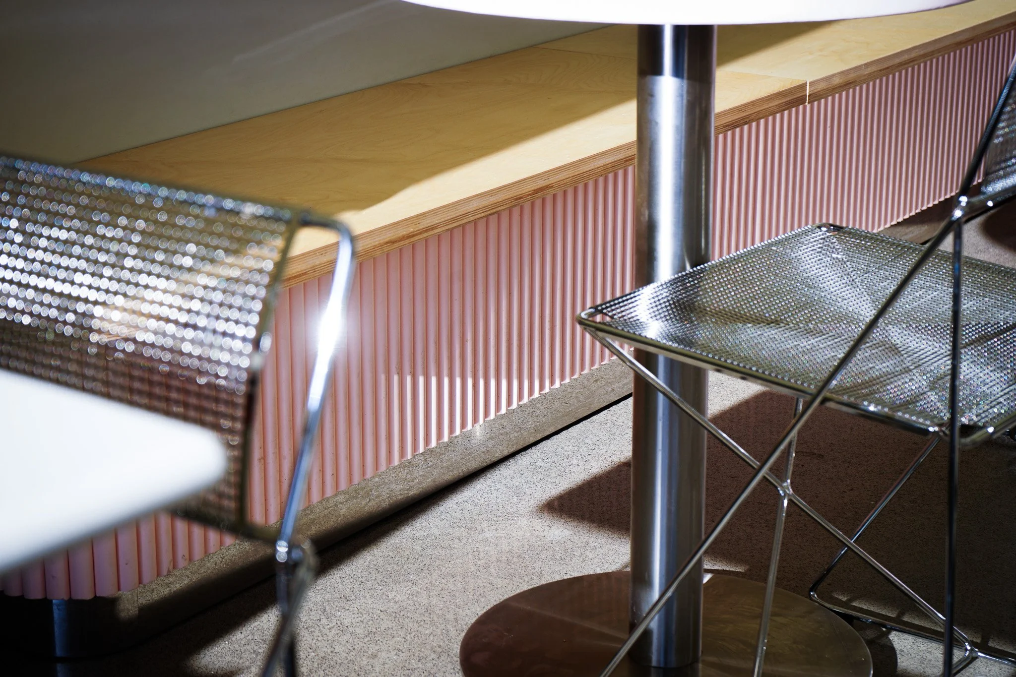 Close-up of metal mesh chairs with black frames around a round table with a metal base, opposite a pink ribbed wall and a beige counter surface.
