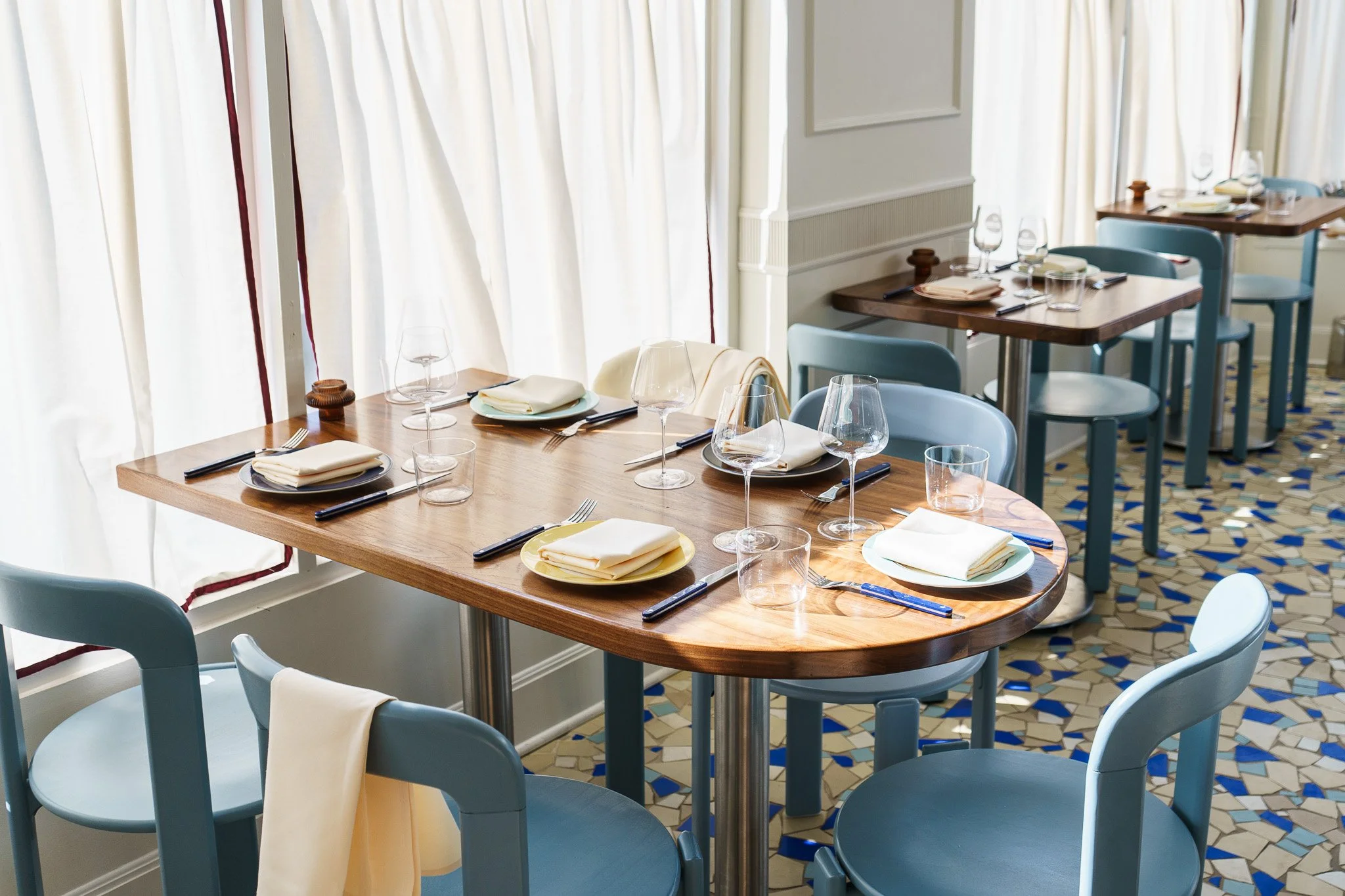 Dining area with wooden tables set with wine glasses, plates, napkins, and cutlery, surrounded by light blue chairs, white curtains, and a patterned tiled floor.