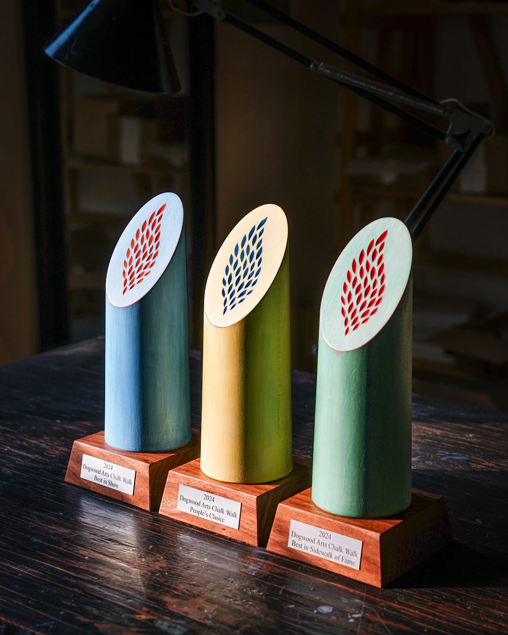 Three colorful wooden trophies with oval shapes and leaf-like designs on top, placed on wooden bases with labels, displayed on a dark wooden surface.
