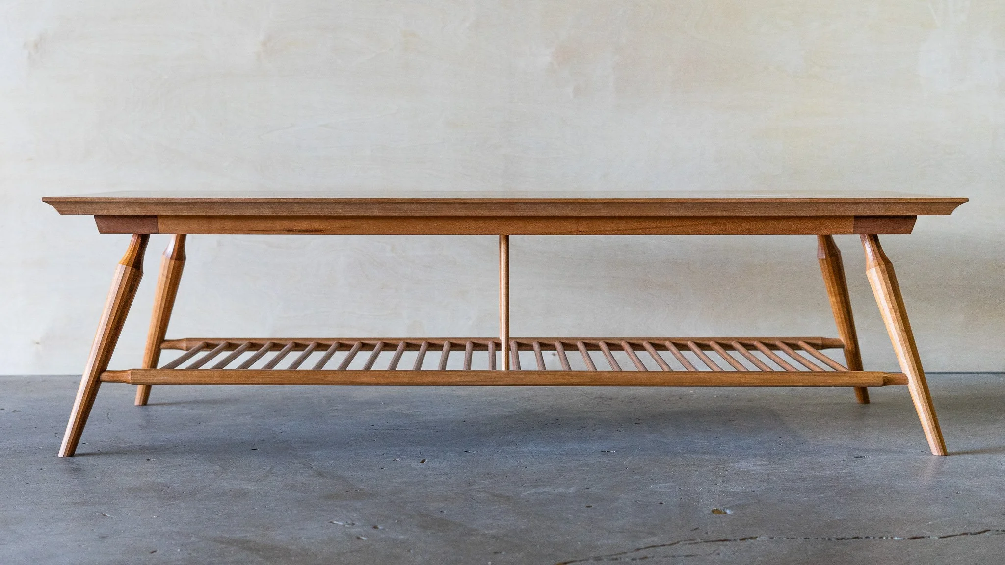 A cherry wooden coffee table with a slatted lower shelf, set against a plain light-colored wall.