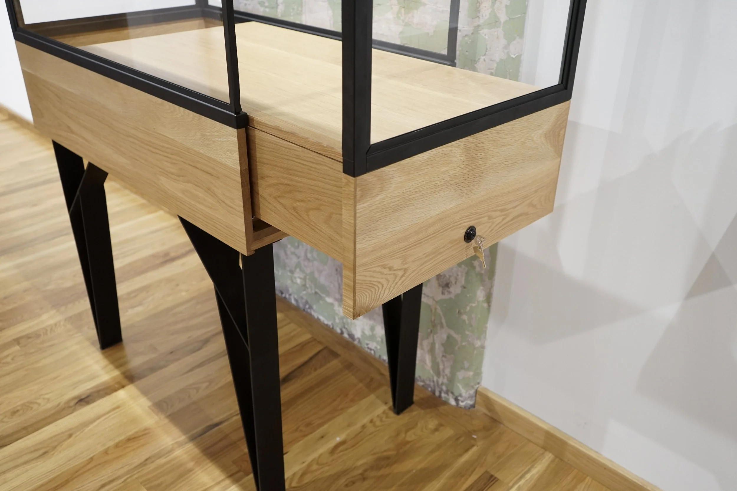 Wooden display case with a glass top and black metal frame, standing on black angled legs, positioned against a partially damaged wall with peeling green paint, on a wooden floor.