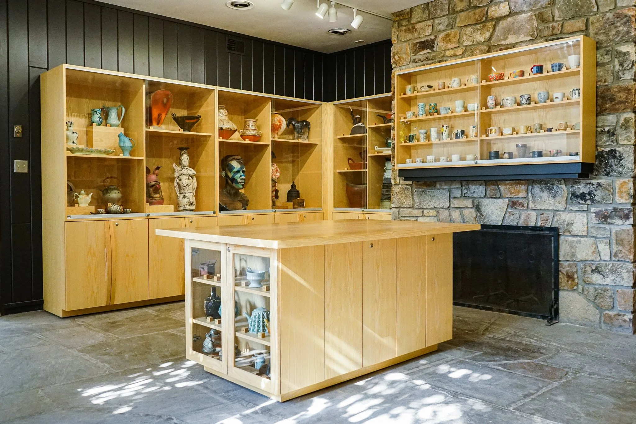 Display of various ceramics and pottery on wooden shelves in a gallery setting with stone and dark wood panel walls.