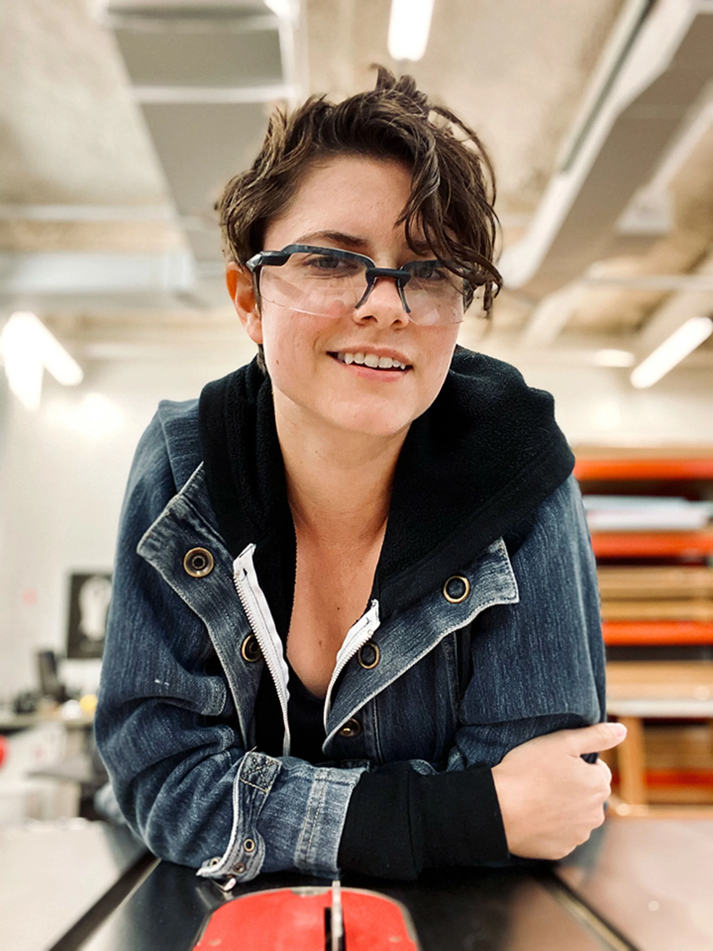 A woman with short, wavy brown hair and glasses smiling at the camera. She is wearing a denim jacket and a black top, sitting at a table in a room with bookshelves and lighting fixtures in the background.