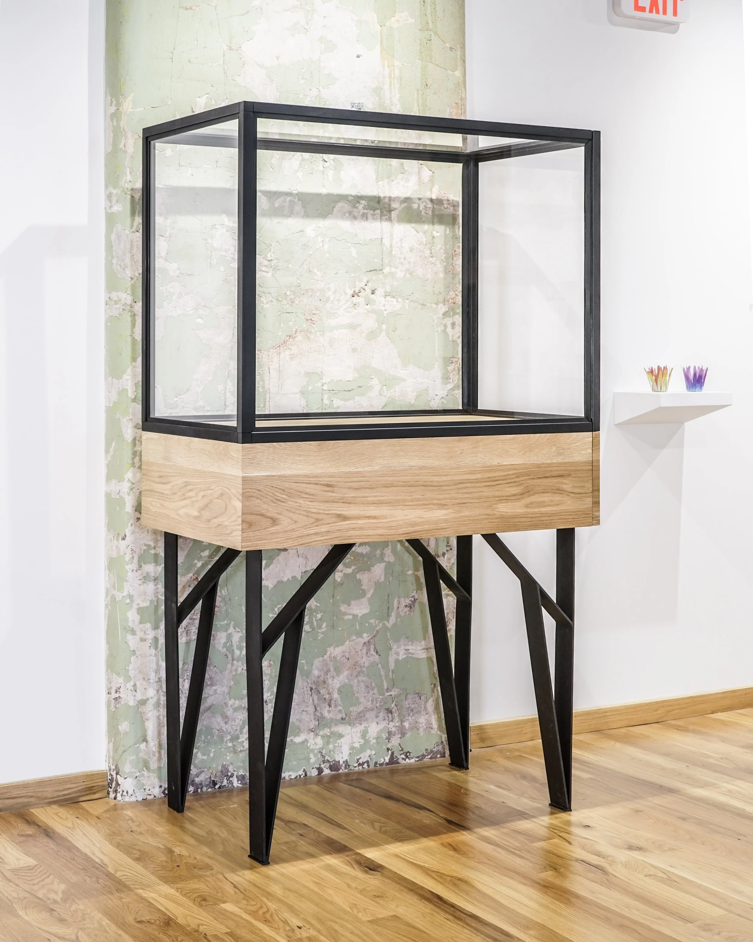 An empty glass display case with black metal frame and wooden base standing on black metal legs in front of a distressed wall with peeling pastel green paint, next to a white wall with a small white shelf holding colorful paper flowers.