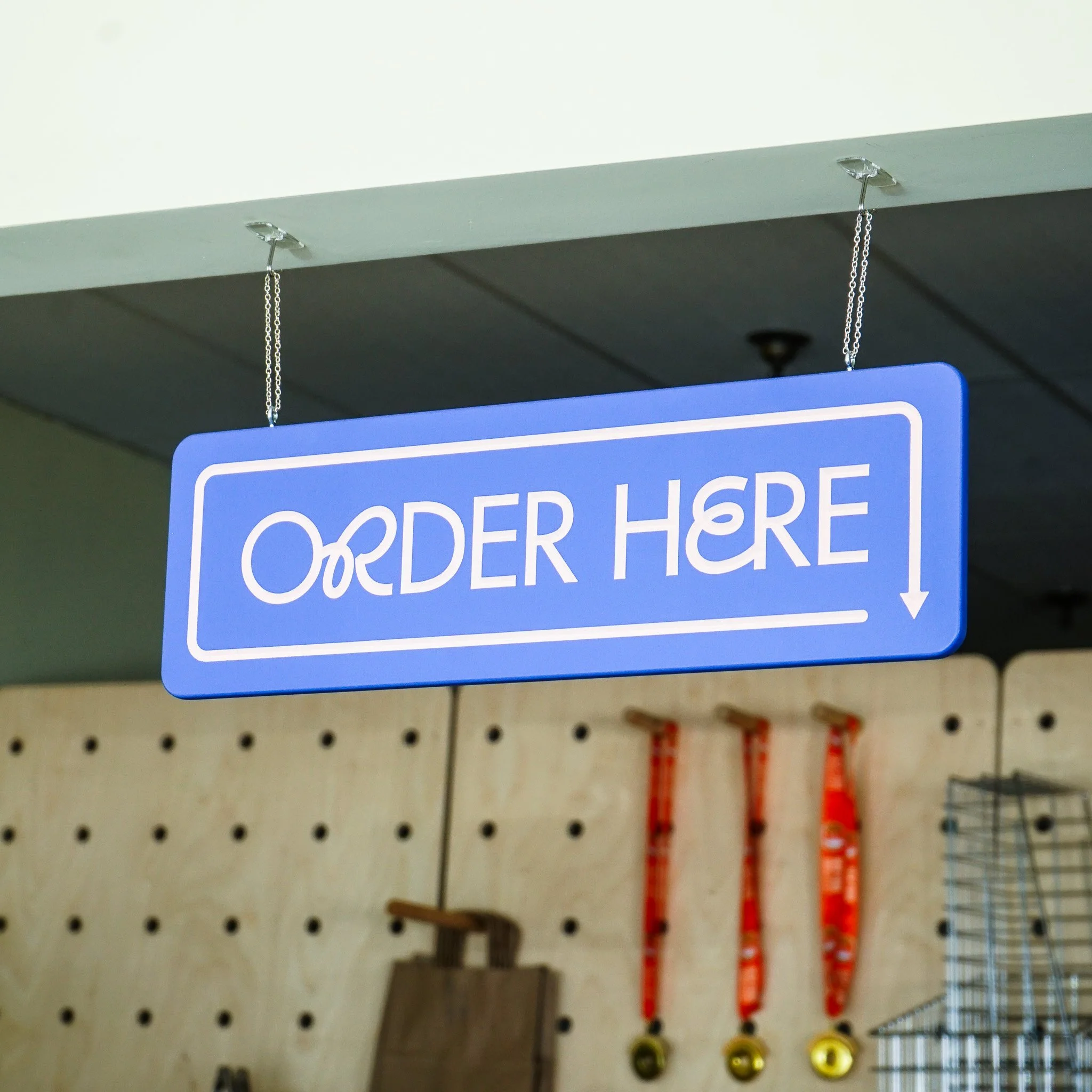 A close-up of a restaurant sign hanging from the ceiling with a metal hook, carved with the words "Order Here".