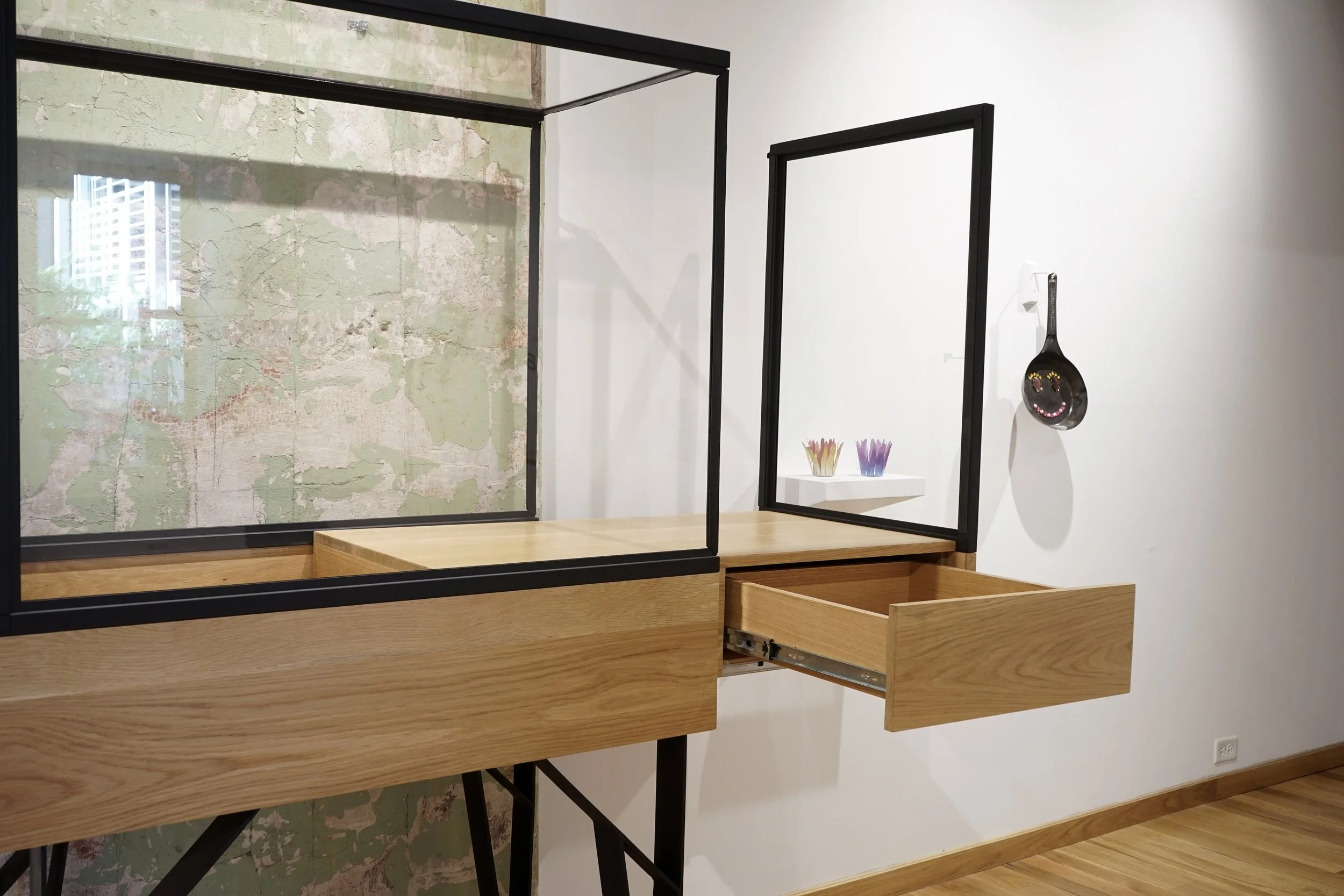 An artistic display features glass panels with black frames on wooden tables, some of which are open drawers. Behind the display, a white wall holds small colorful ceramic containers and a spoon-shaped wall decor with a smiling face.