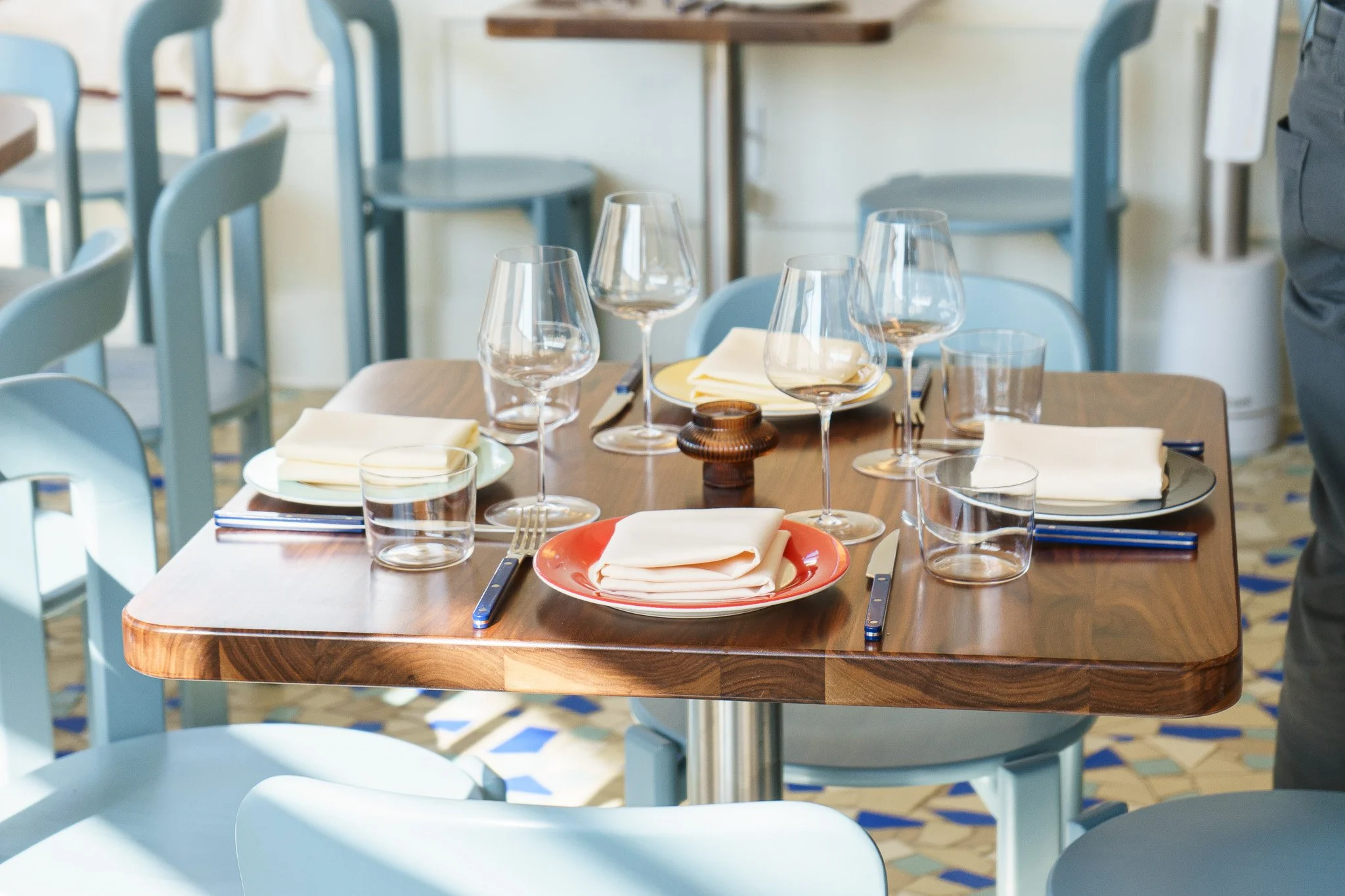 A restaurant table set for a meal, with wine glasses, water glasses, plates with napkins, cutlery, and a small brown candle holder, in a room with blue chairs and a patterned tile floor.