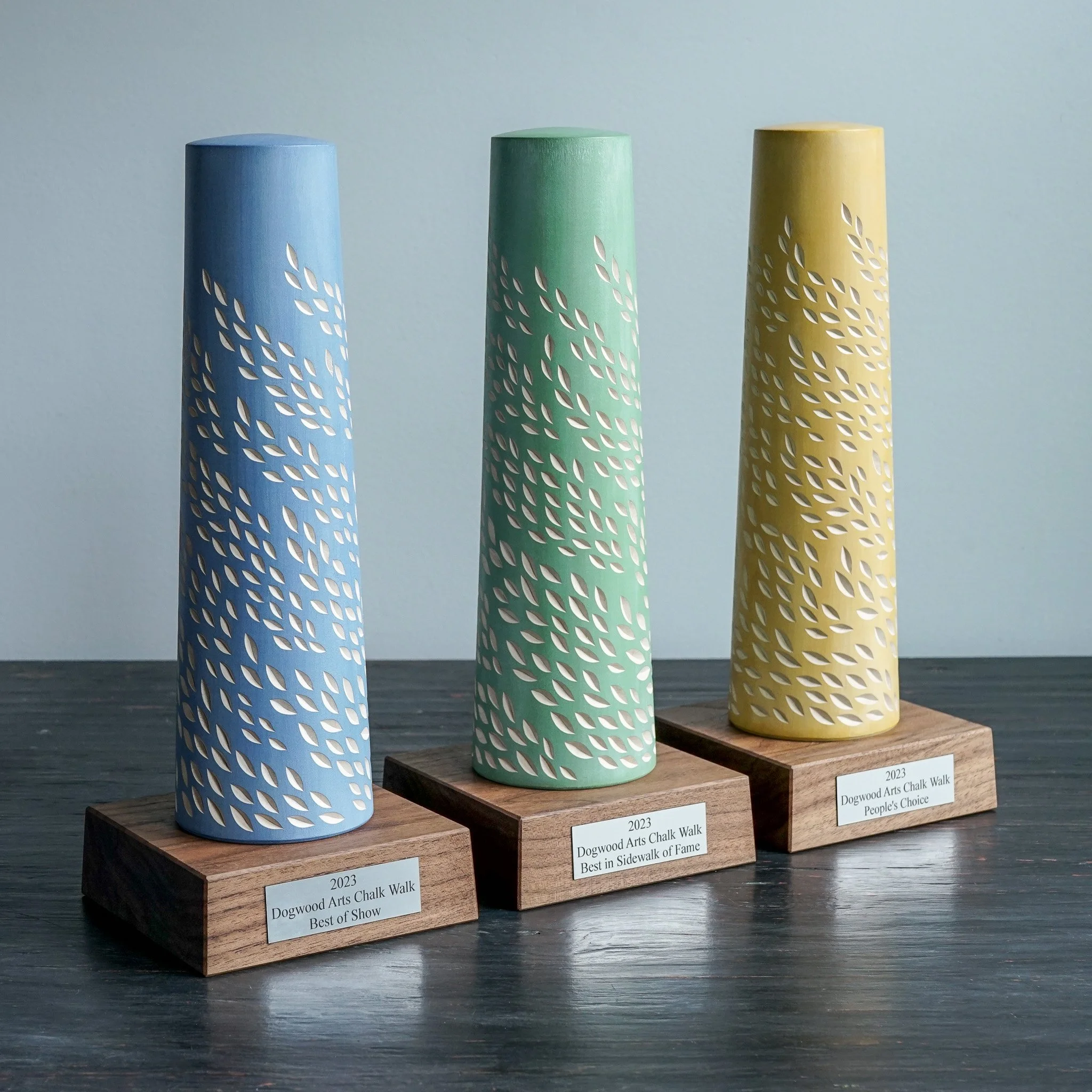 Three cylindrical ceramic vases painted in pastel colors (blue, green, yellow) with cutout leaf patterns, displayed on wooden bases with labels indicating they are award-winning chalk walk artwork from 2023, part of a sidewalk art show.