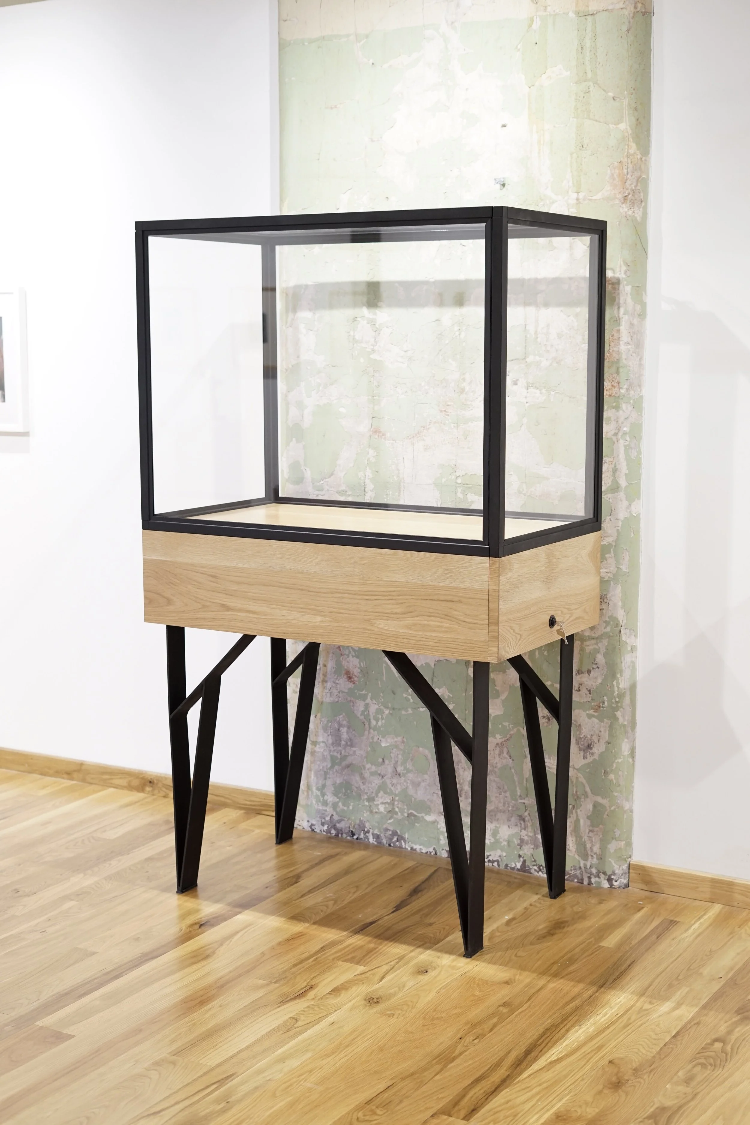A display case with a black frame and glass sides on a wooden stand, set against a textured wall with peeling paint and a white wall.