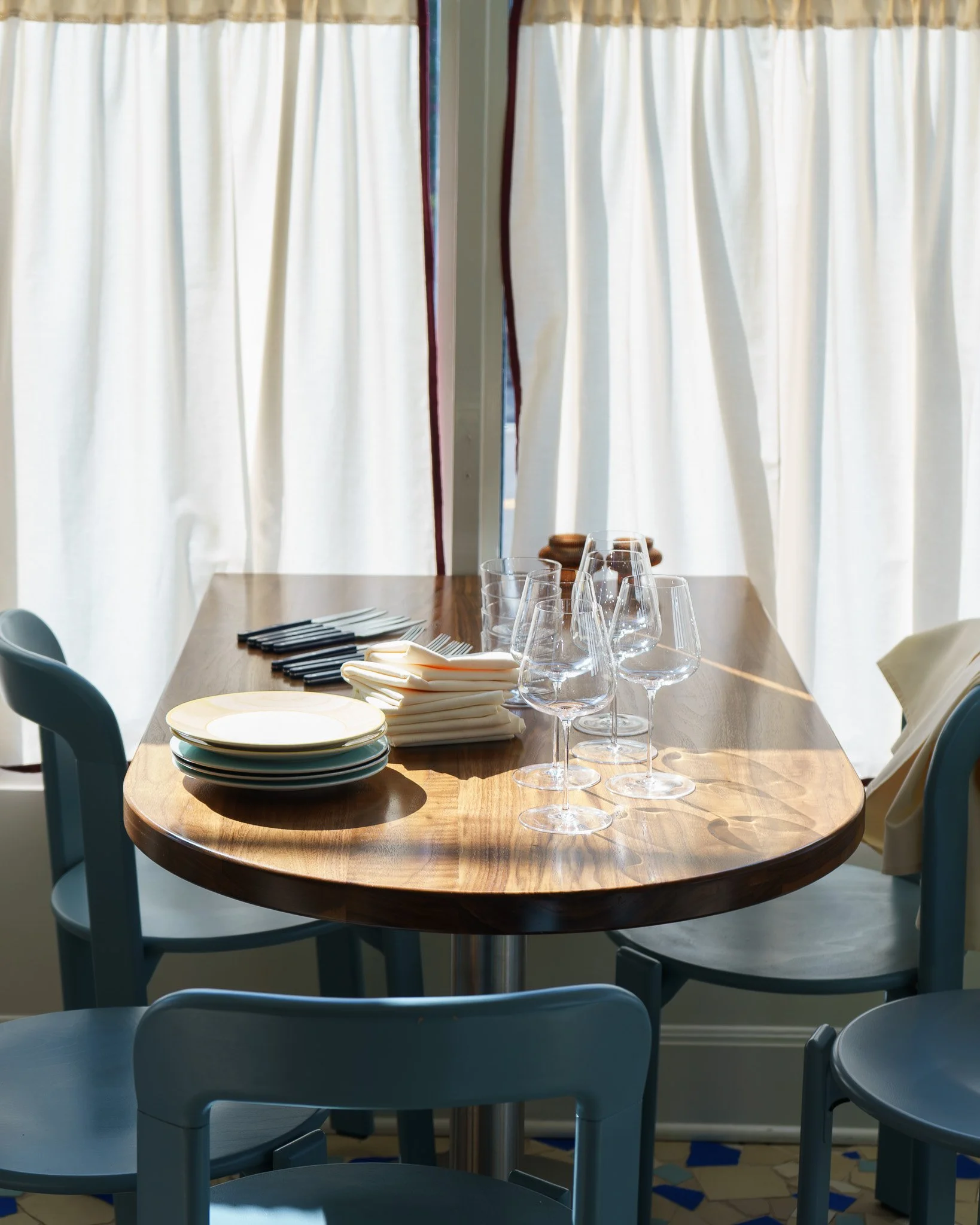 A dining table set with plates, napkins, and wine glasses, with sunlight coming through white curtains in the background.