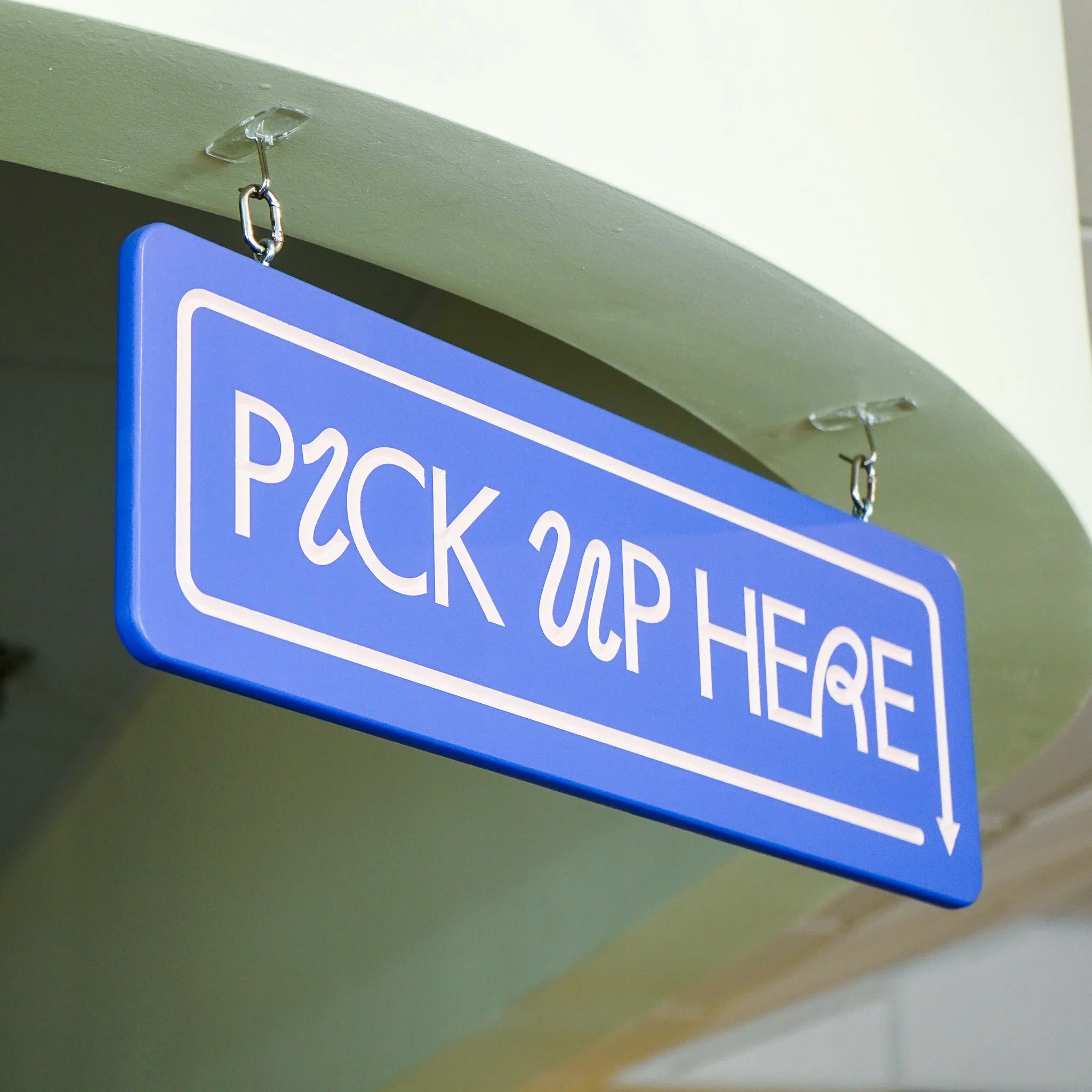 A close-up of a restaurant sign hanging from the ceiling with a metal hook, carved with the words "Pick Up Here".