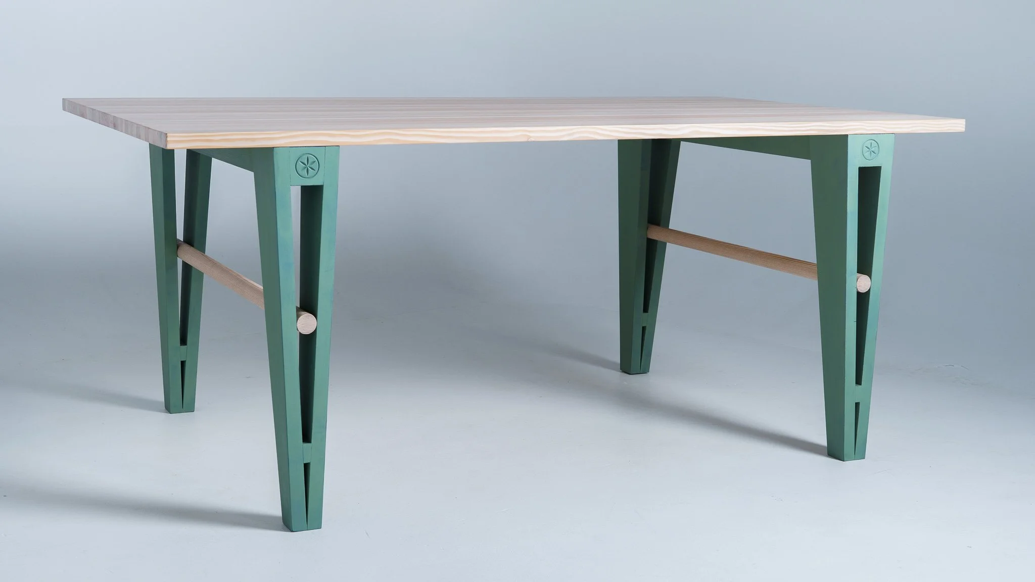 A wooden dining table with green legs and light-colored top, set against a light blue background.