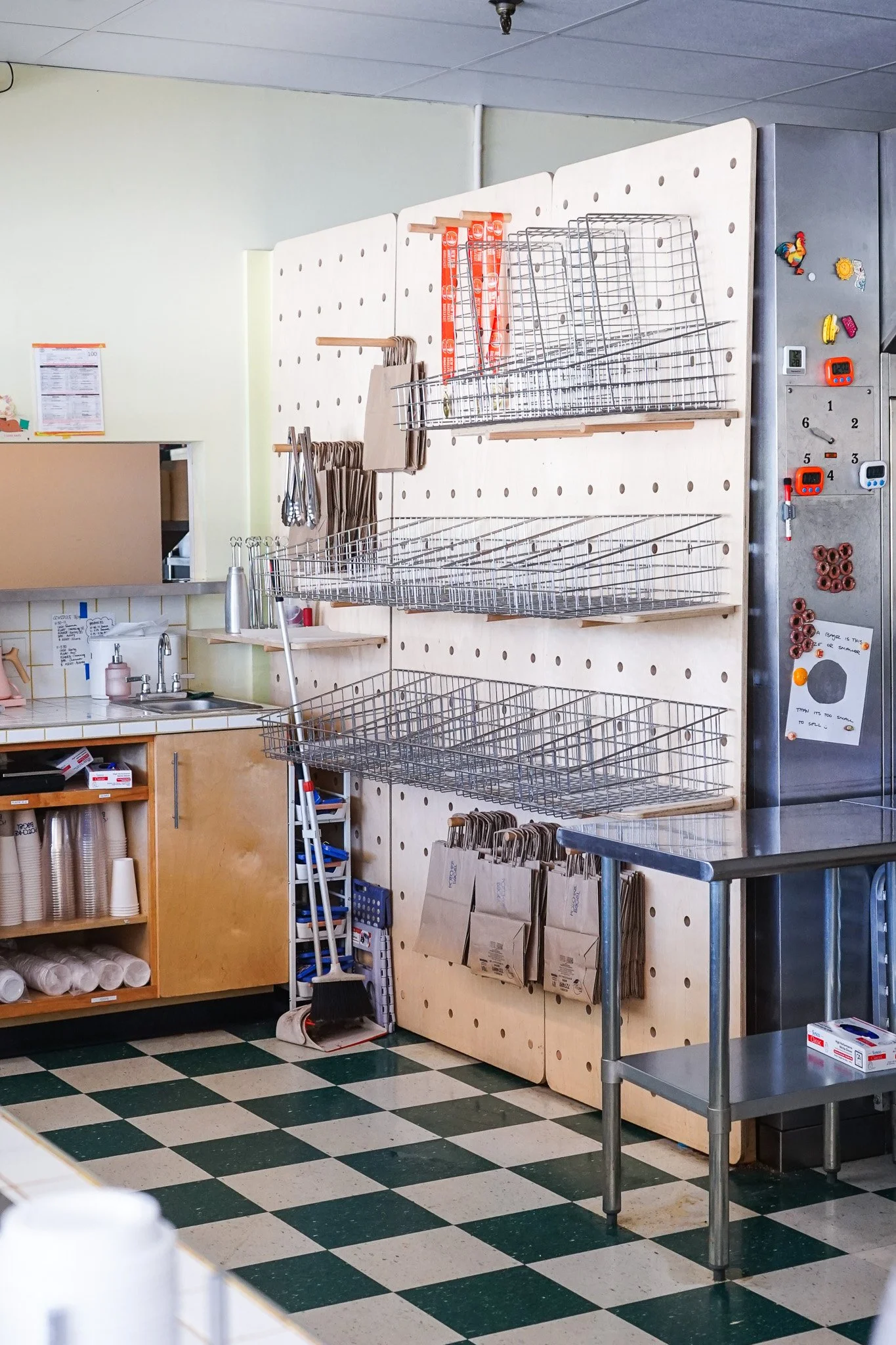 A section of a store or restaurant with a pegboard wall containing metal baskets, brown paper bags, and hanging utensils. There are paper bags hanging on hooks, a small sink area with cleaning supplies, and a stainless steel prep table. The floor has