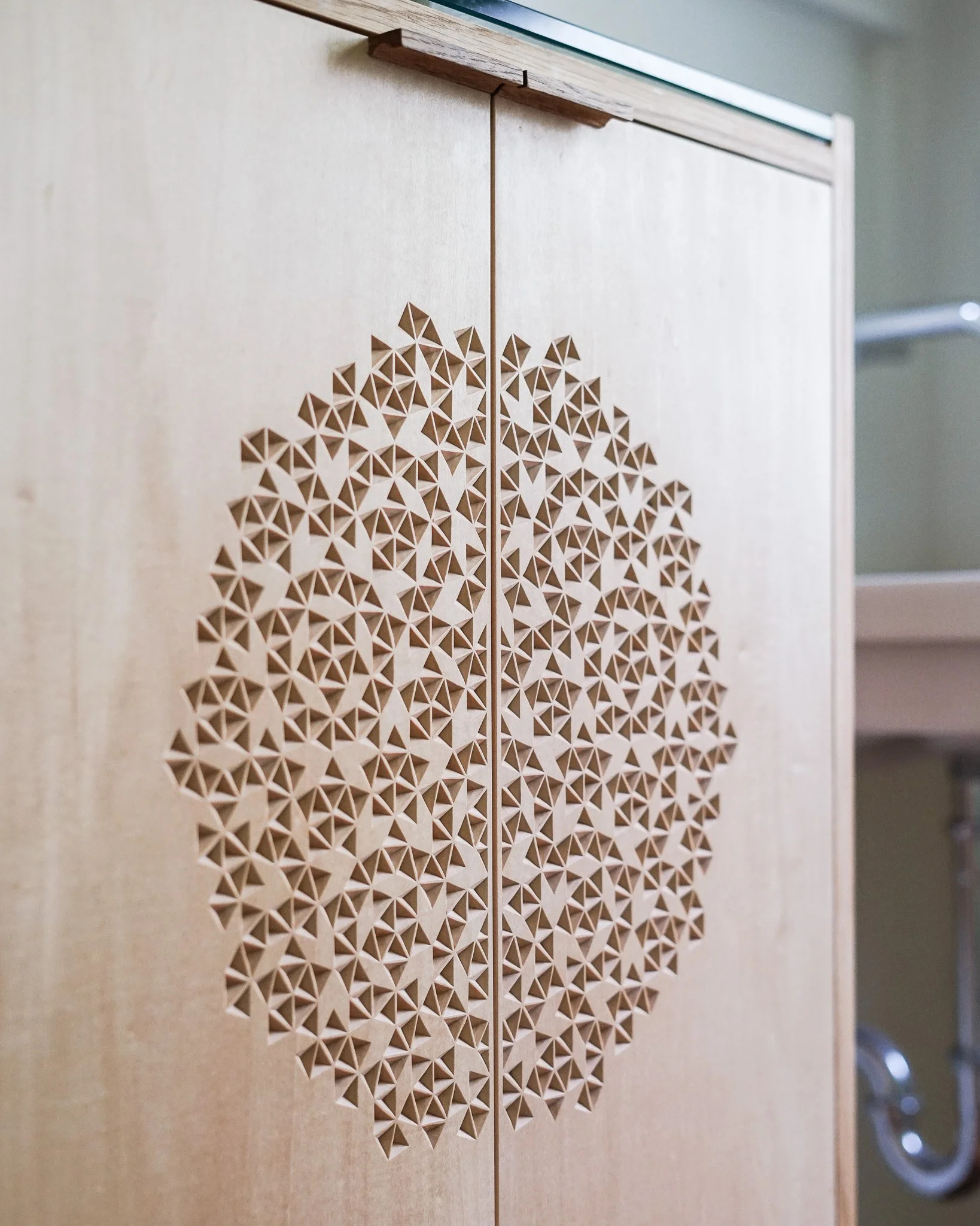 A detail image of a wooden bathroom vanity cabinet with hand carved door panels in front of a beige walll near a mirror and bathroom sink.