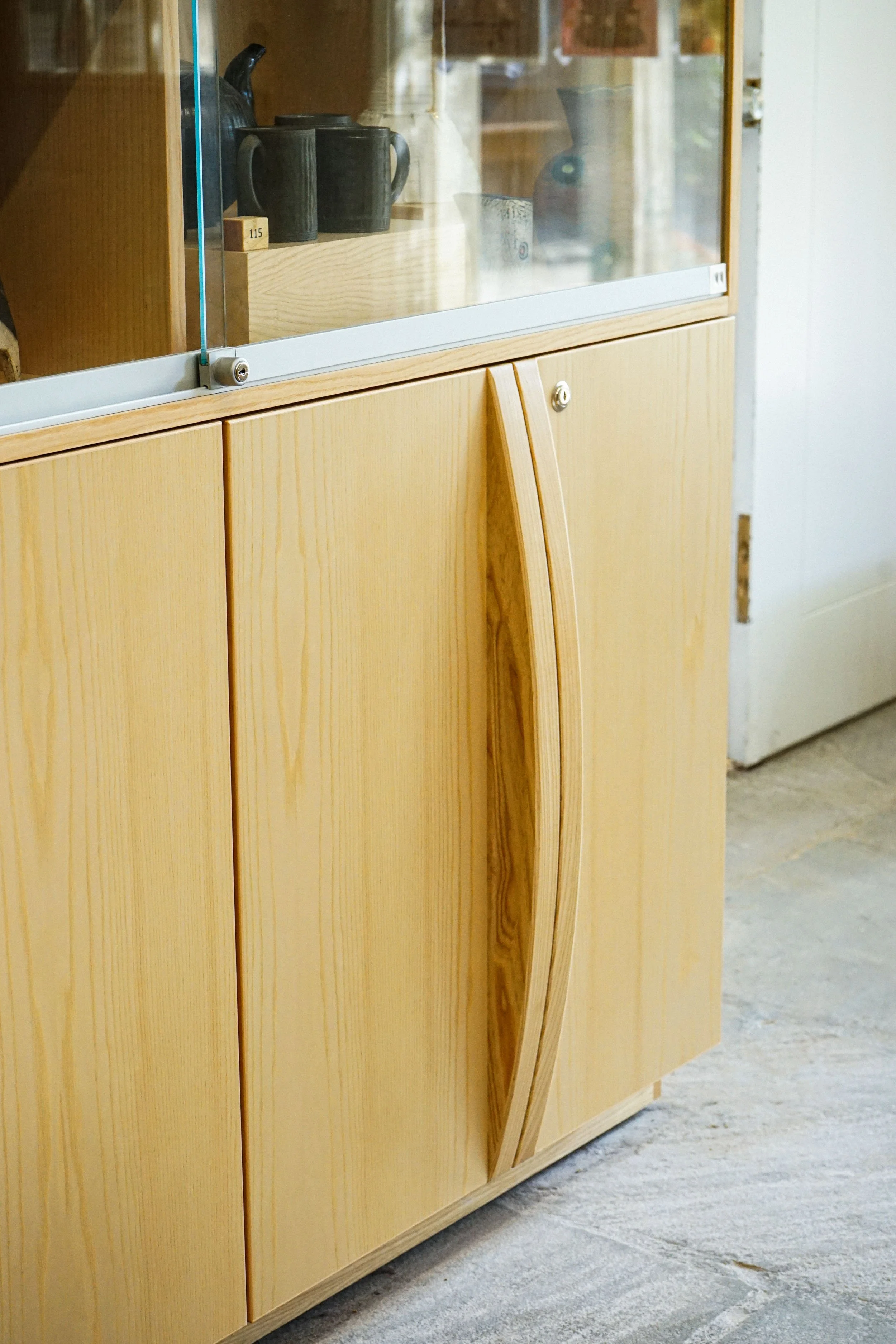 Light wood cabinet with glass-front doors displaying dark ceramic mugs, a stoneware vase, and a small block labeled '115' inside.