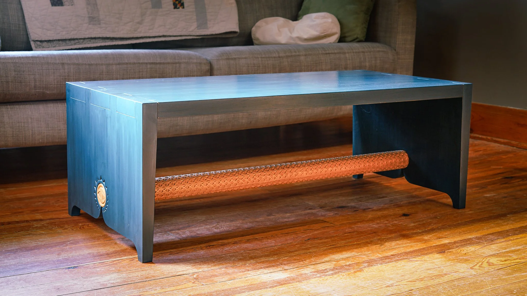Blue rectangular coffee table with a hand carved wooden medal on one side, placed on wooden floor in front of a brown sofa with pillows and a quilted blanket.